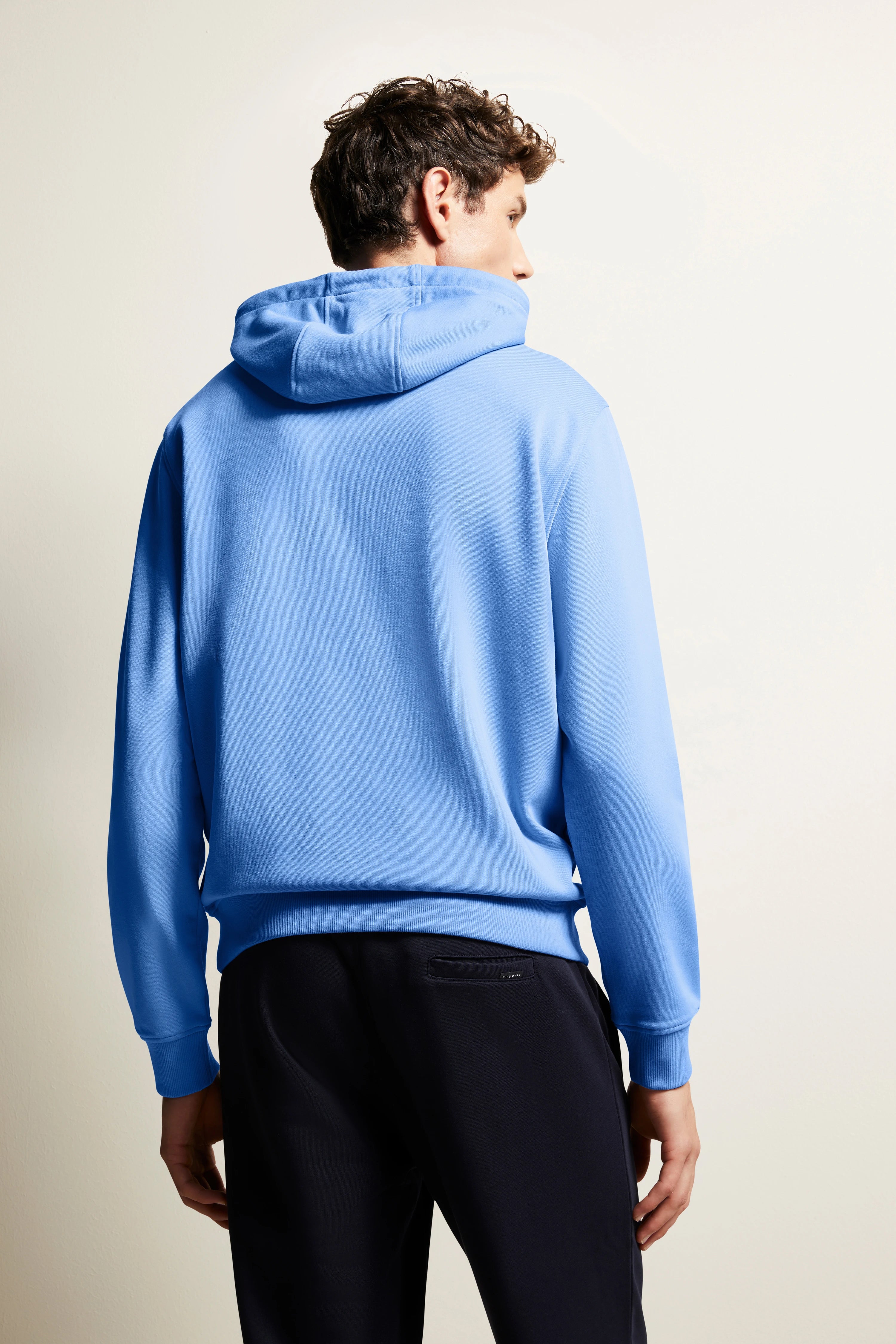 bugatti Modelbild: Mann in blauem Hoodie und dunkler Hose, Rückenansicht, entspanntes Posing.