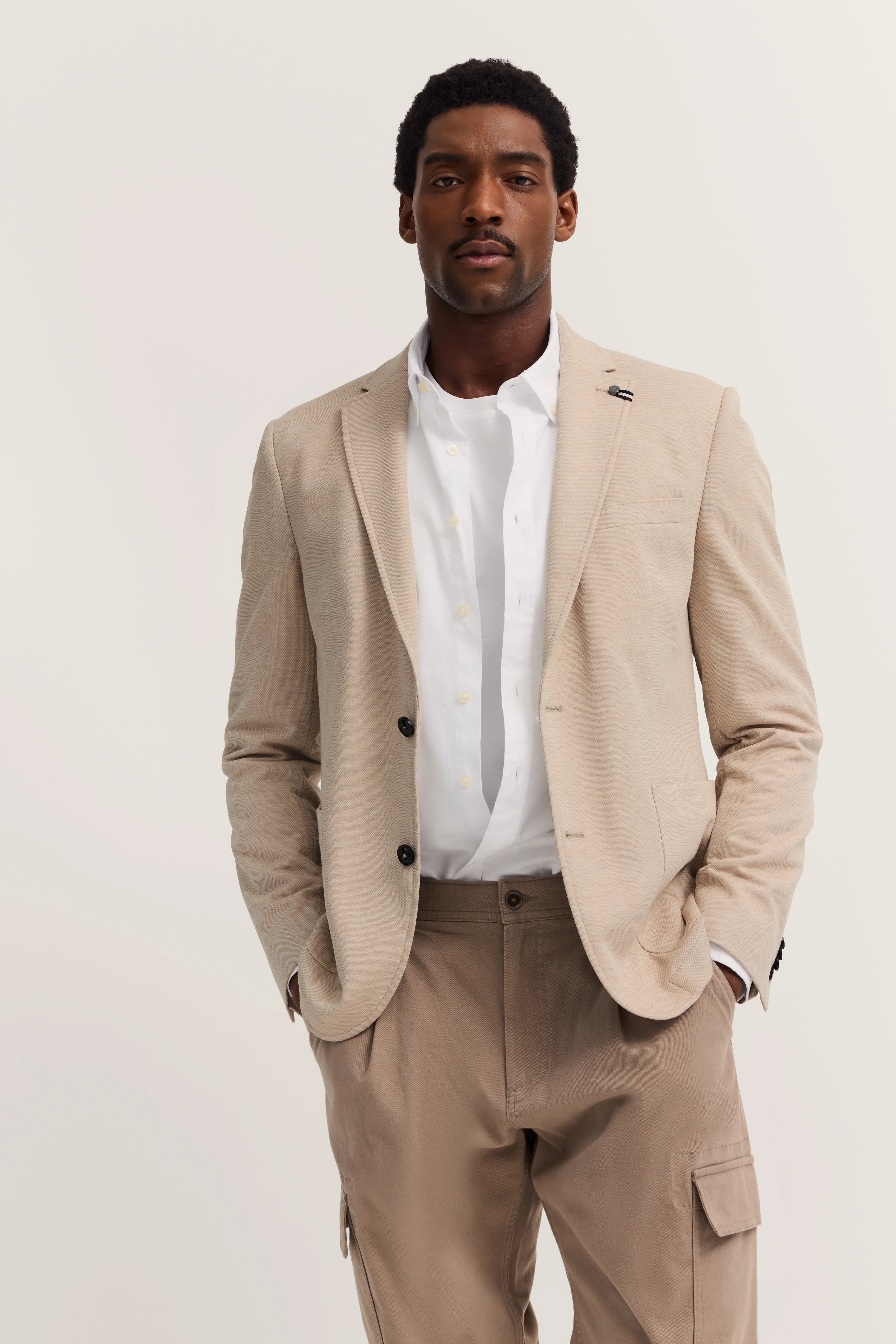 Elegantes Model in beigem bugatti Blazer und weißem Hemd. Lässige Pose mit Händen in Taschen.