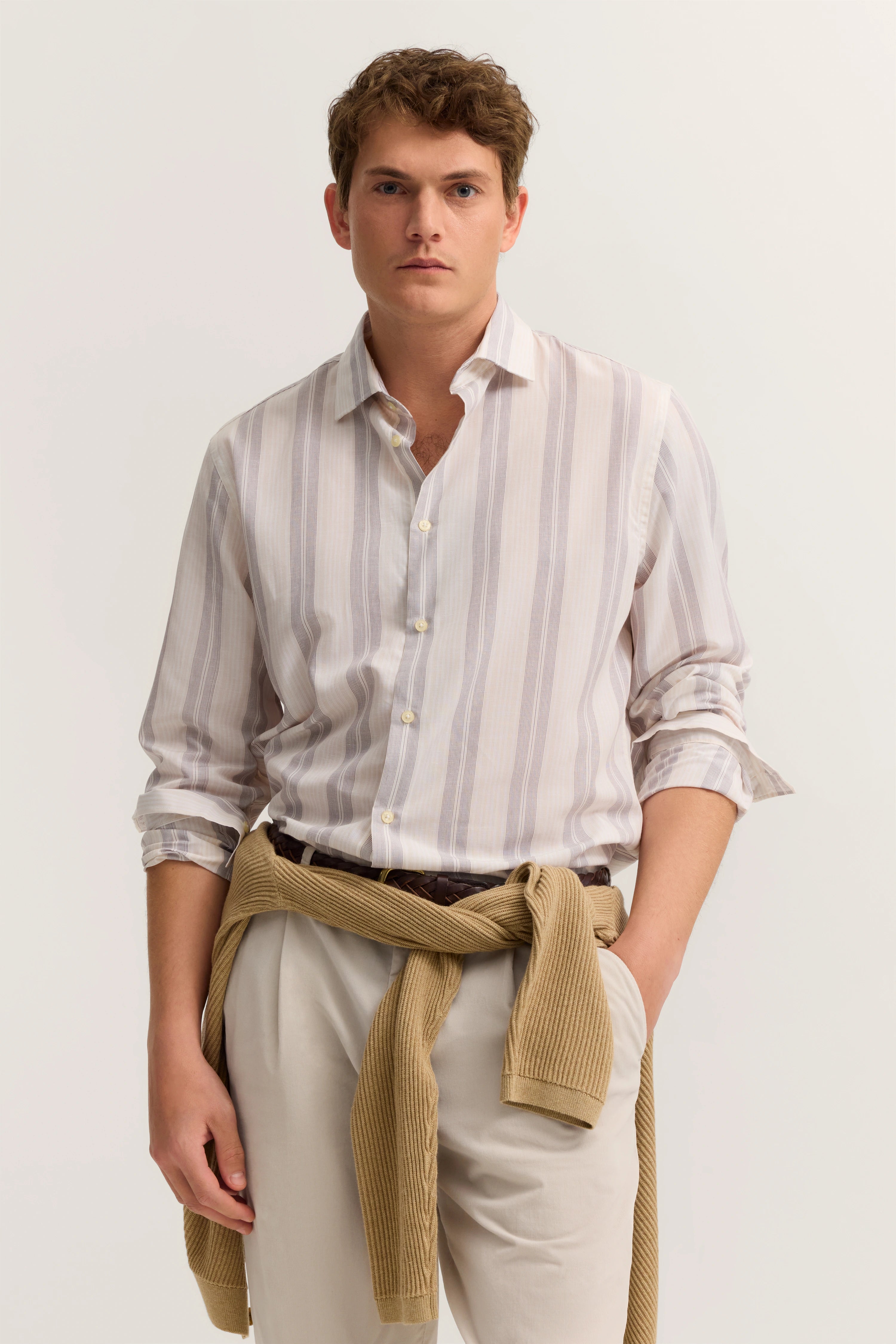 Lässiges Model in gestreiftem Hemd, heller Hose, beige Strickjacke um die Taille, entspannte Pose.