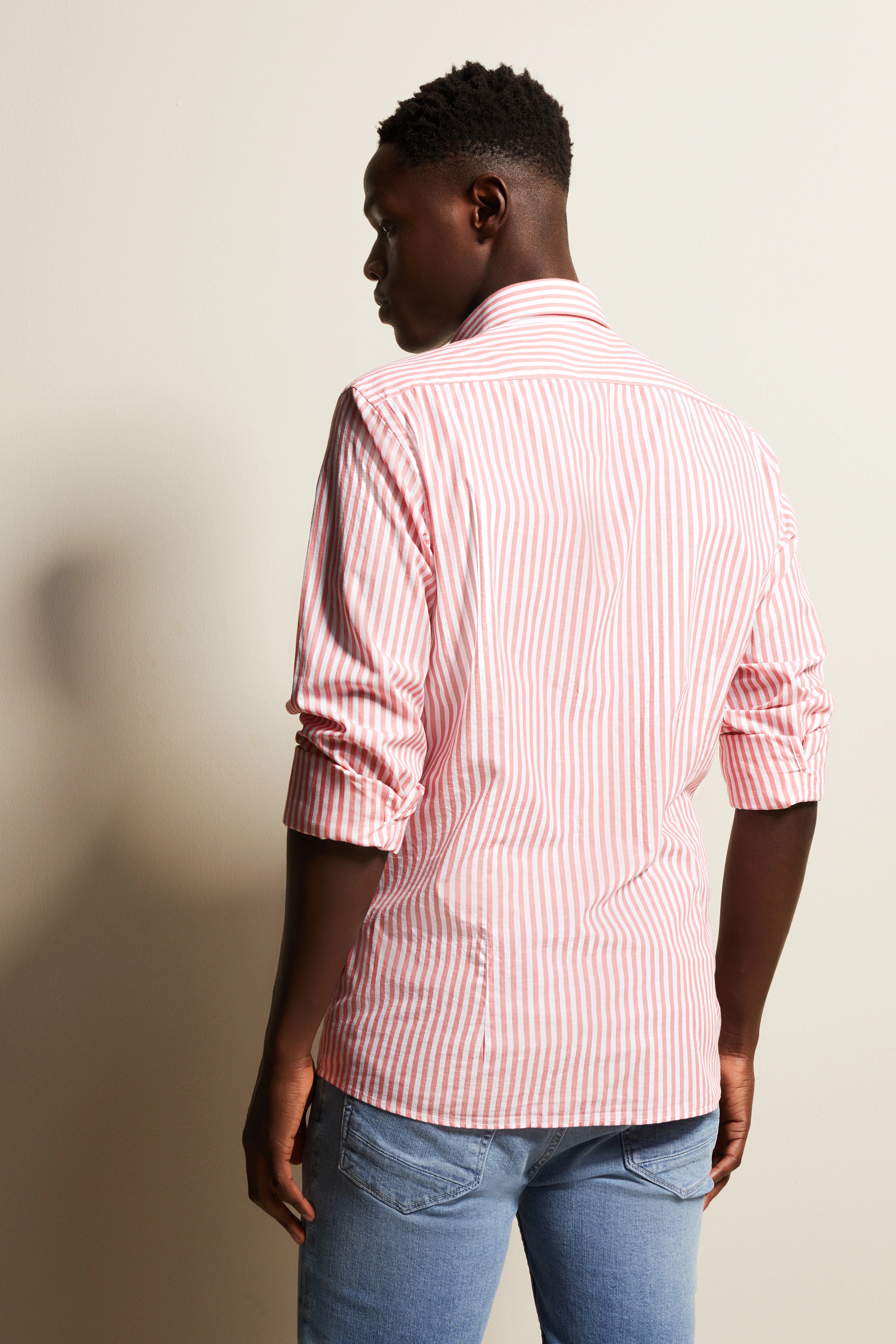 Rear view of model in bugatti striped pink-white shirt and blue jeans, relaxed pose.