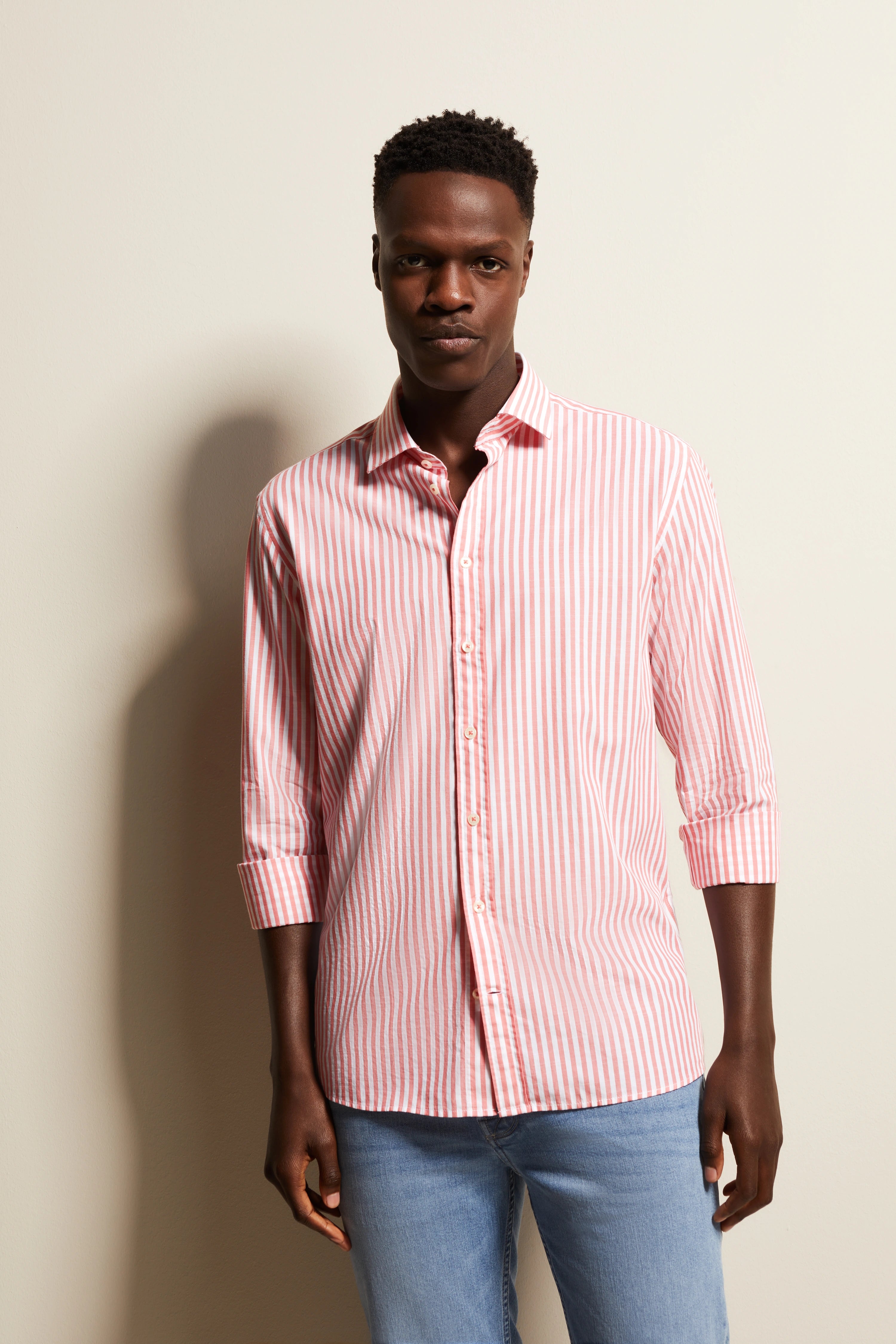 Model in pink-white striped bugatti shirt, casually posing, paired with blue jeans.