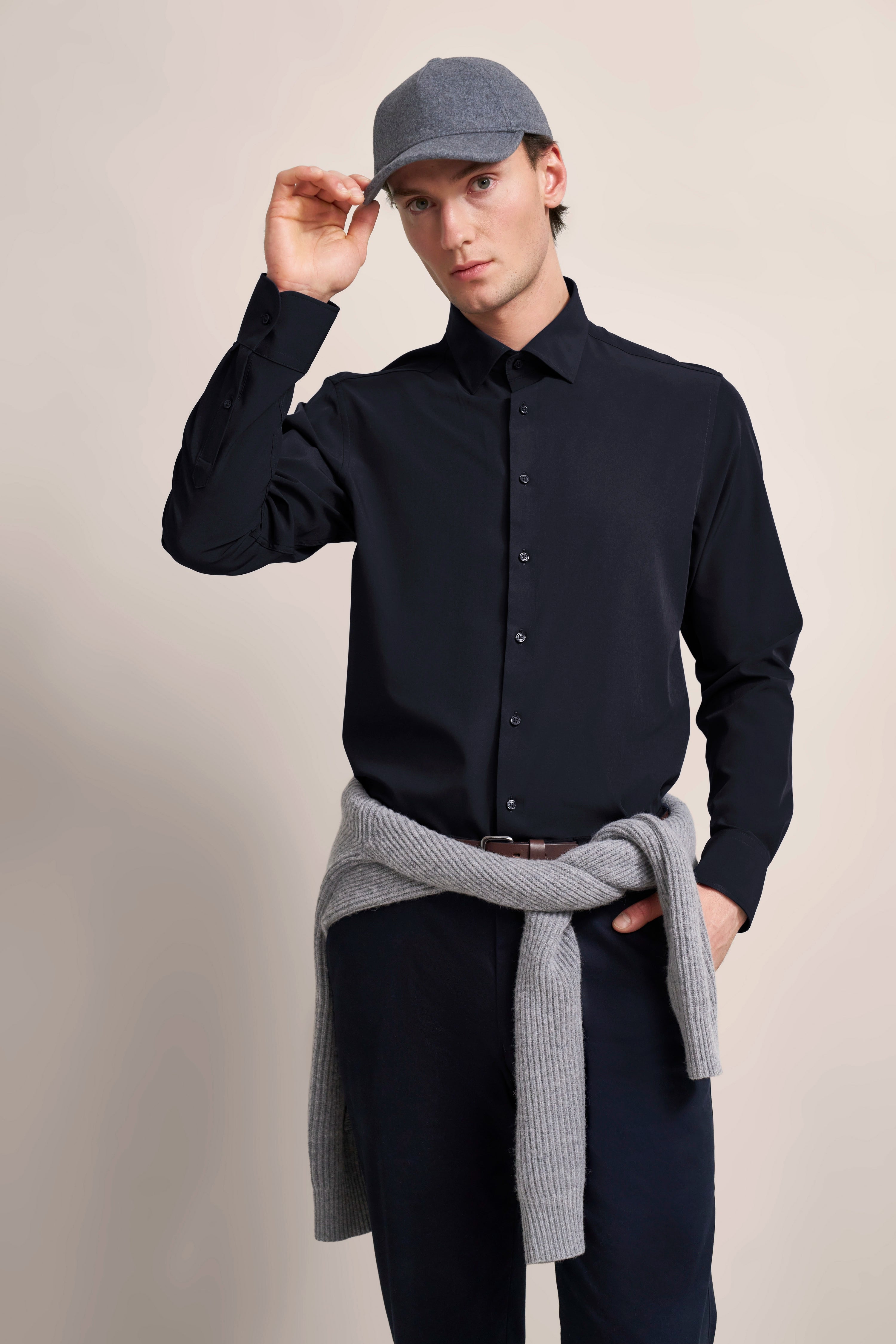 Modelbild: Mann in schwarzem Hemd, grauer Mütze, grauer Pullover um die Hüfte gebunden. Lässige Pose.