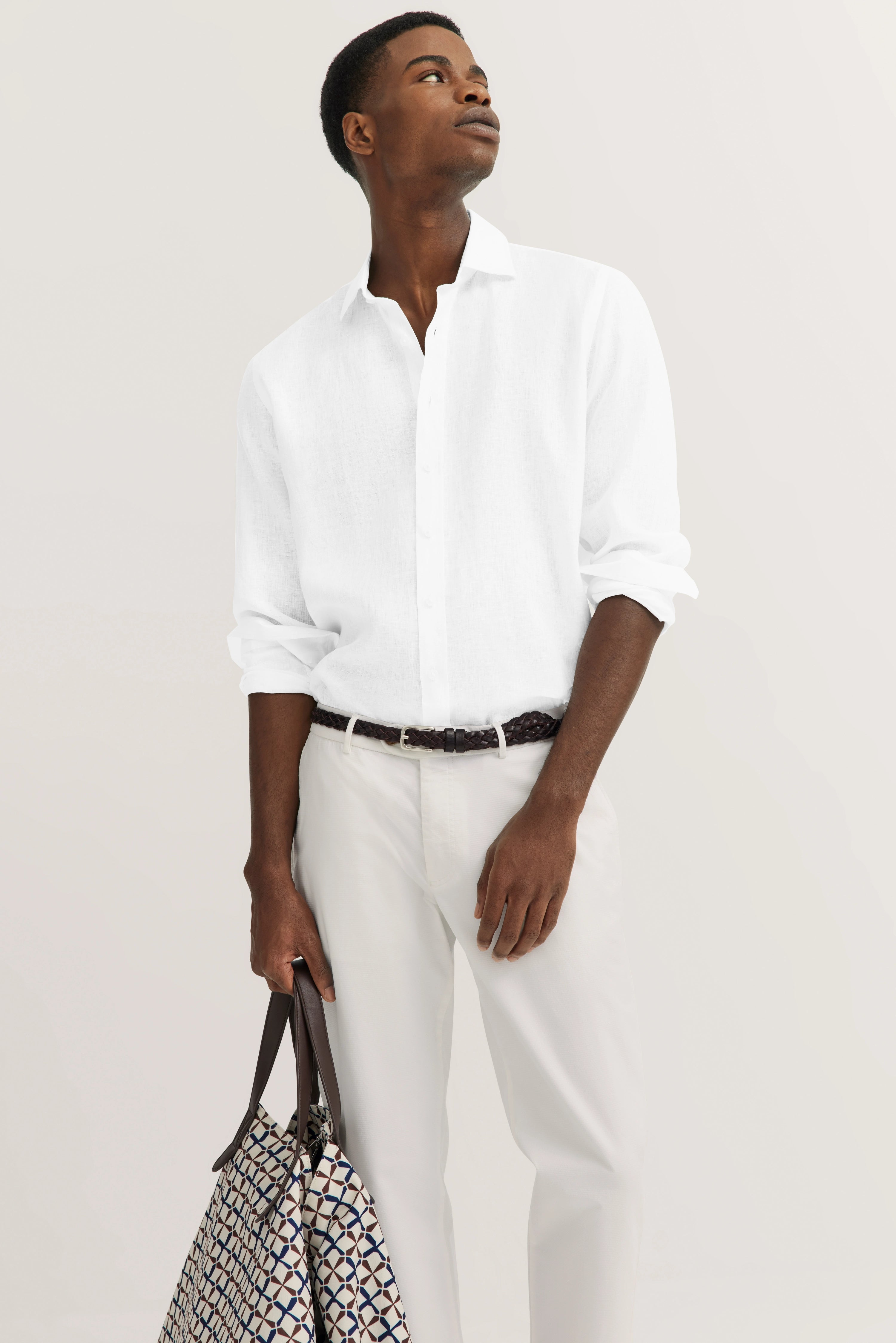 Elegantes bugatti Outfit: Weißes Hemd, weiße Hose, Model mit gemusterter Tasche, entspannter Blick.