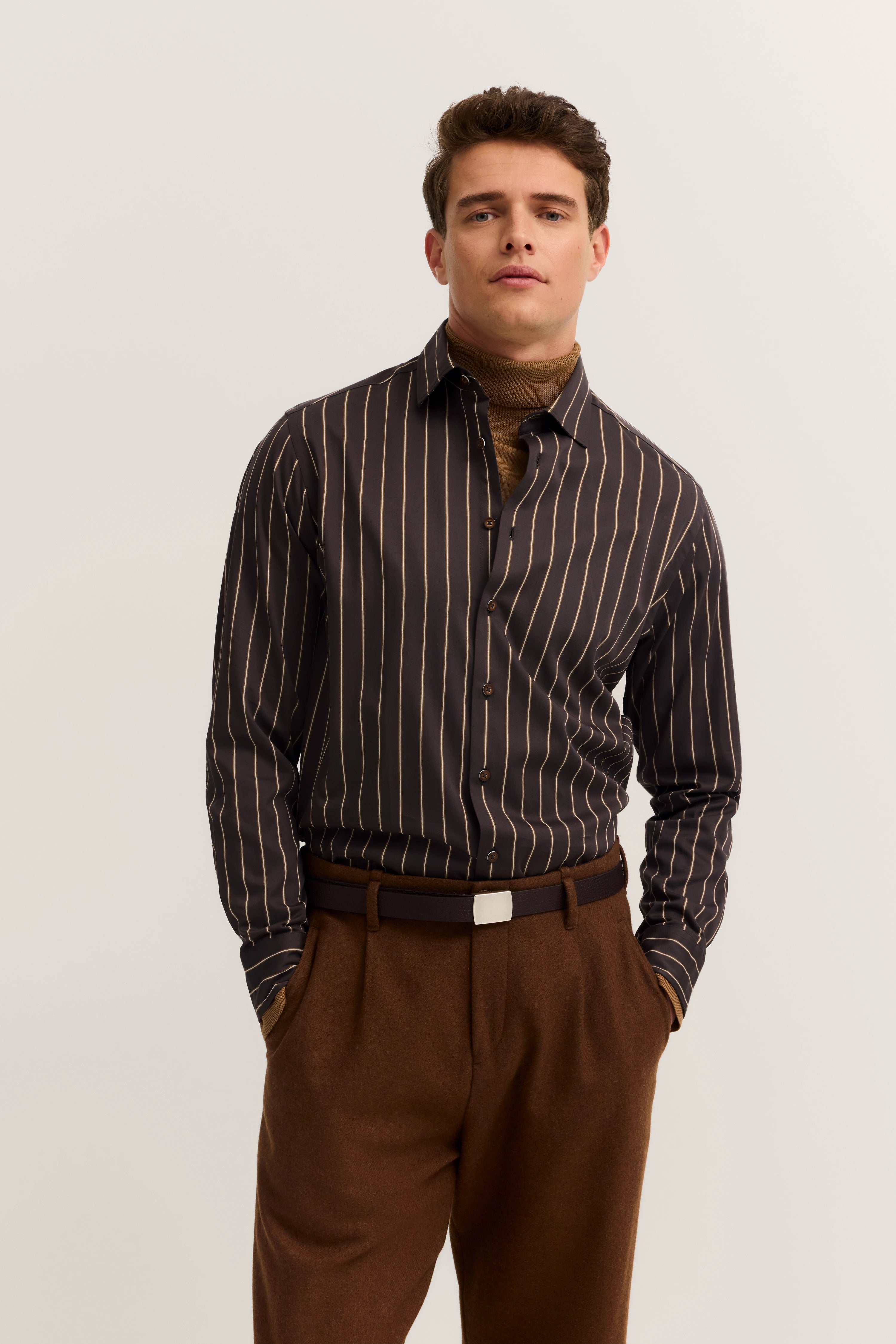Model trägt gestreiftes Hemd, braunen Rollkragenpullover und Hose. Lässige Pose, bugatti Stil.