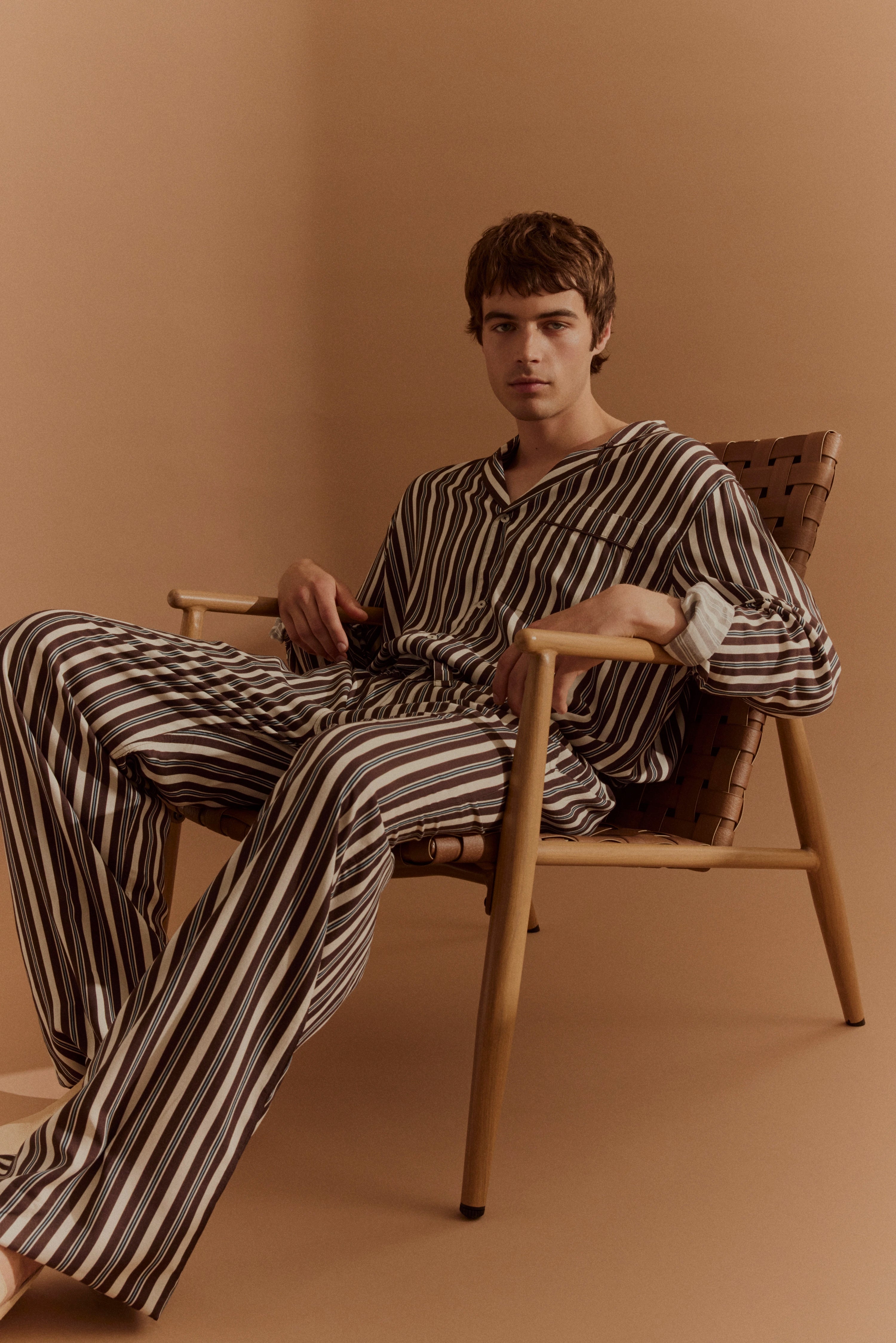 Modell in gestreiftem, braun-beigem Pyjama, entspannt sitzend auf einem Stuhl, minimalistisches Setting.