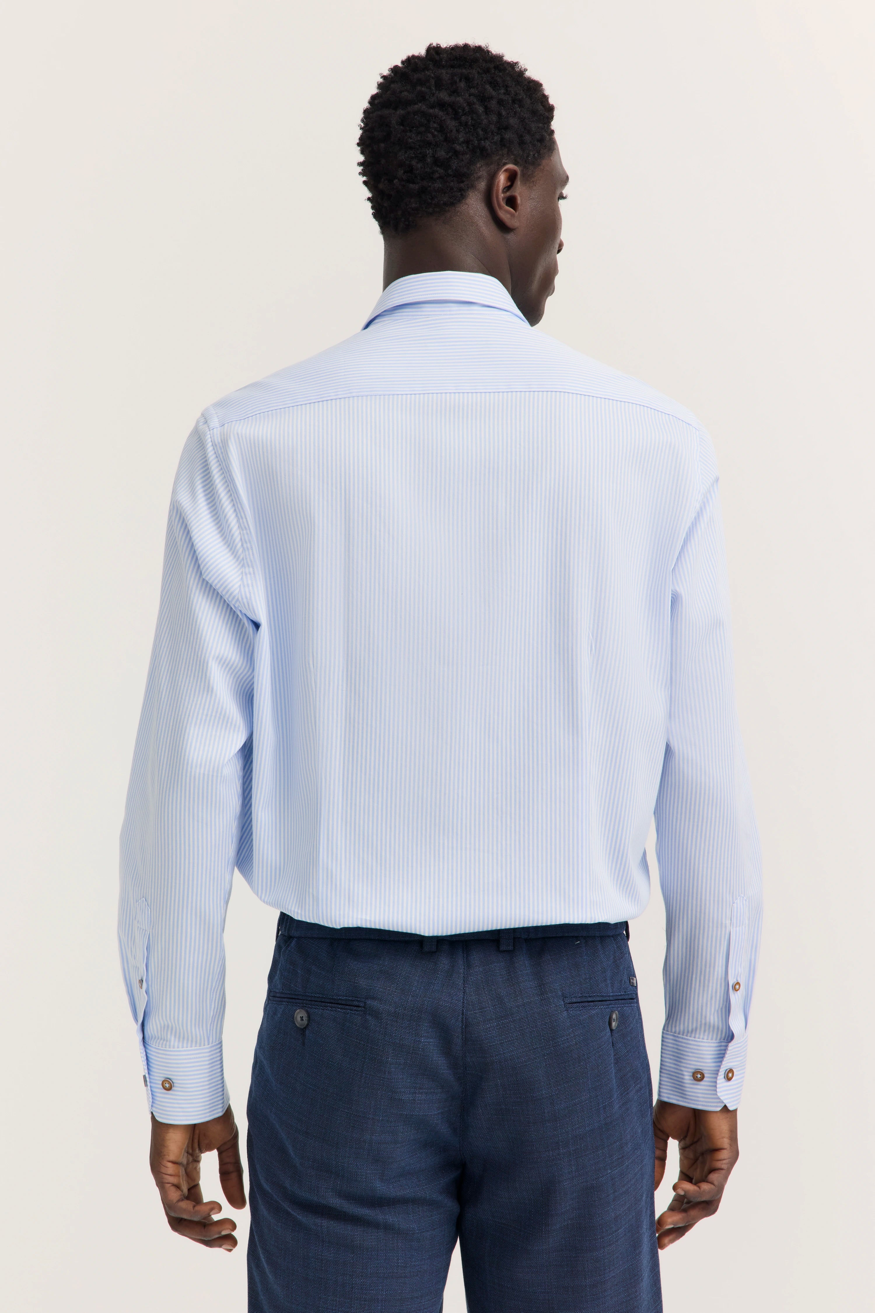 Ein Model trägt ein hellblau gestreiftes Hemd und dunkelblaue Hose. Rückenansicht, entspannte Haltung.