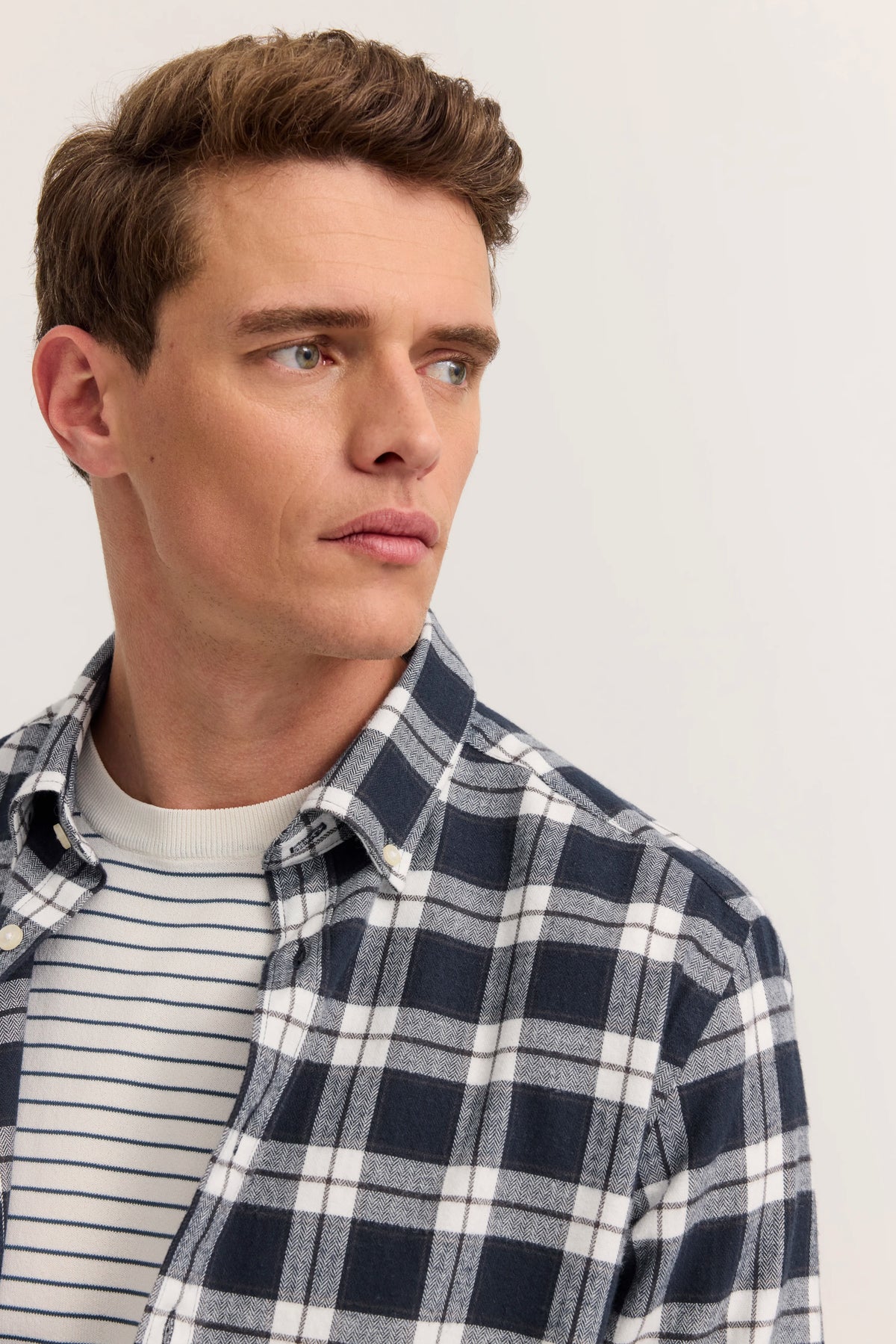 Büstenbild: Model trägt kariertes Hemd in Blau und Weiß über gestreiftem Pullover, schaut seitlich.