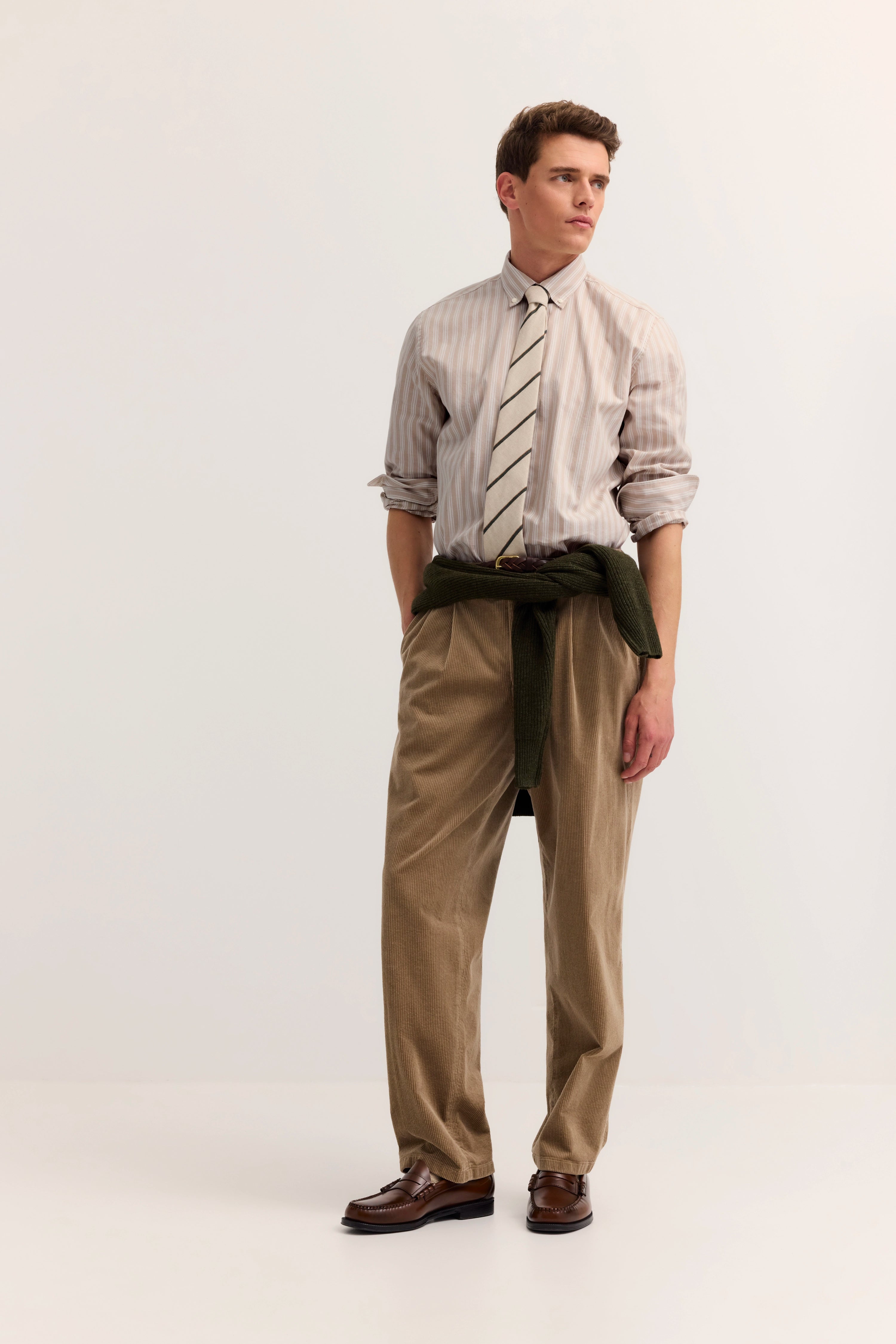 Model in gestreiftem Hemd, Krawatte, beigefarbener Hose, grüner Pullover um die Taille, formell posierend.
