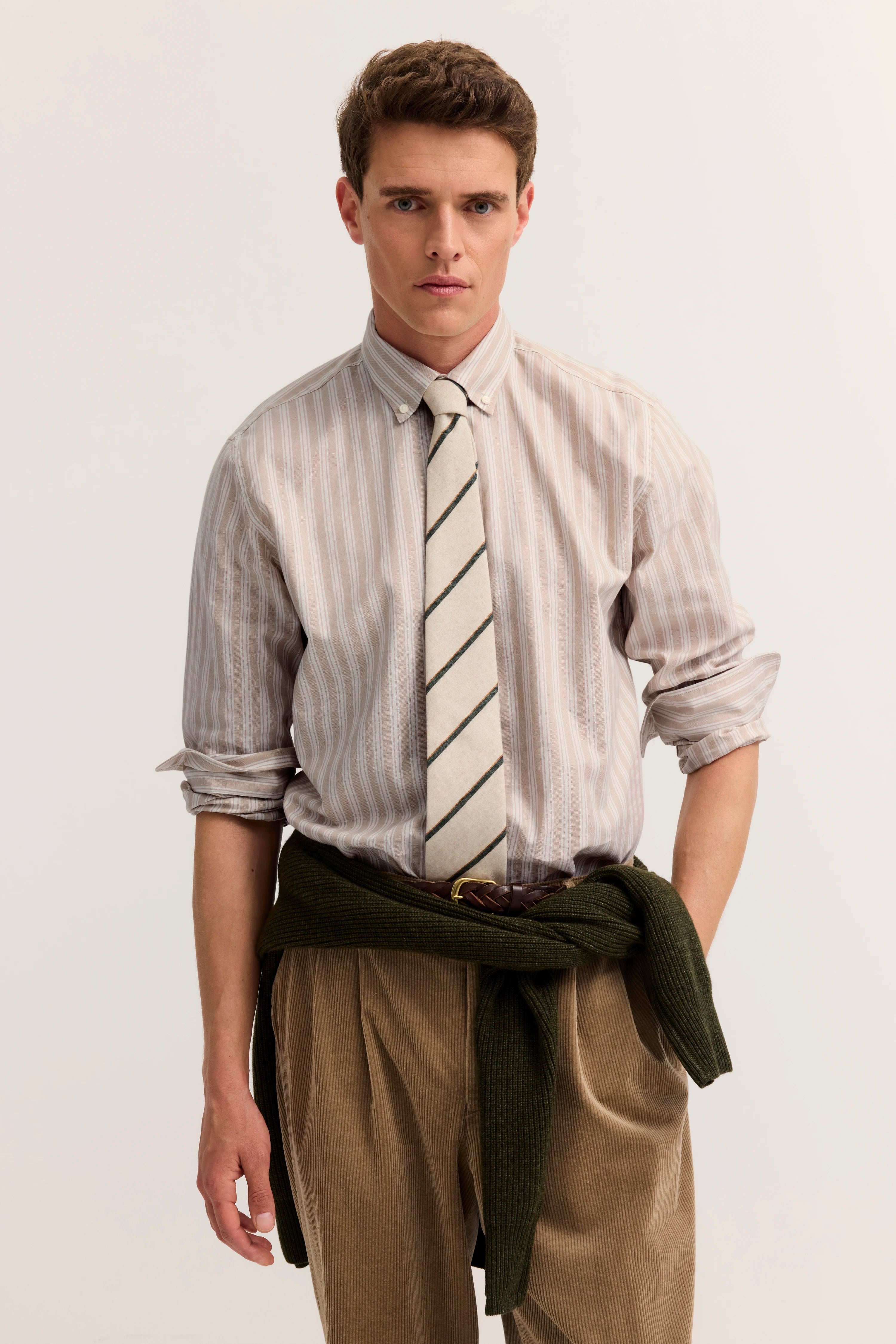 Streifenhemd mit Krawatte, Cordhose, Pullover um Taille gebunden, Modelbild, entspannte Pose.