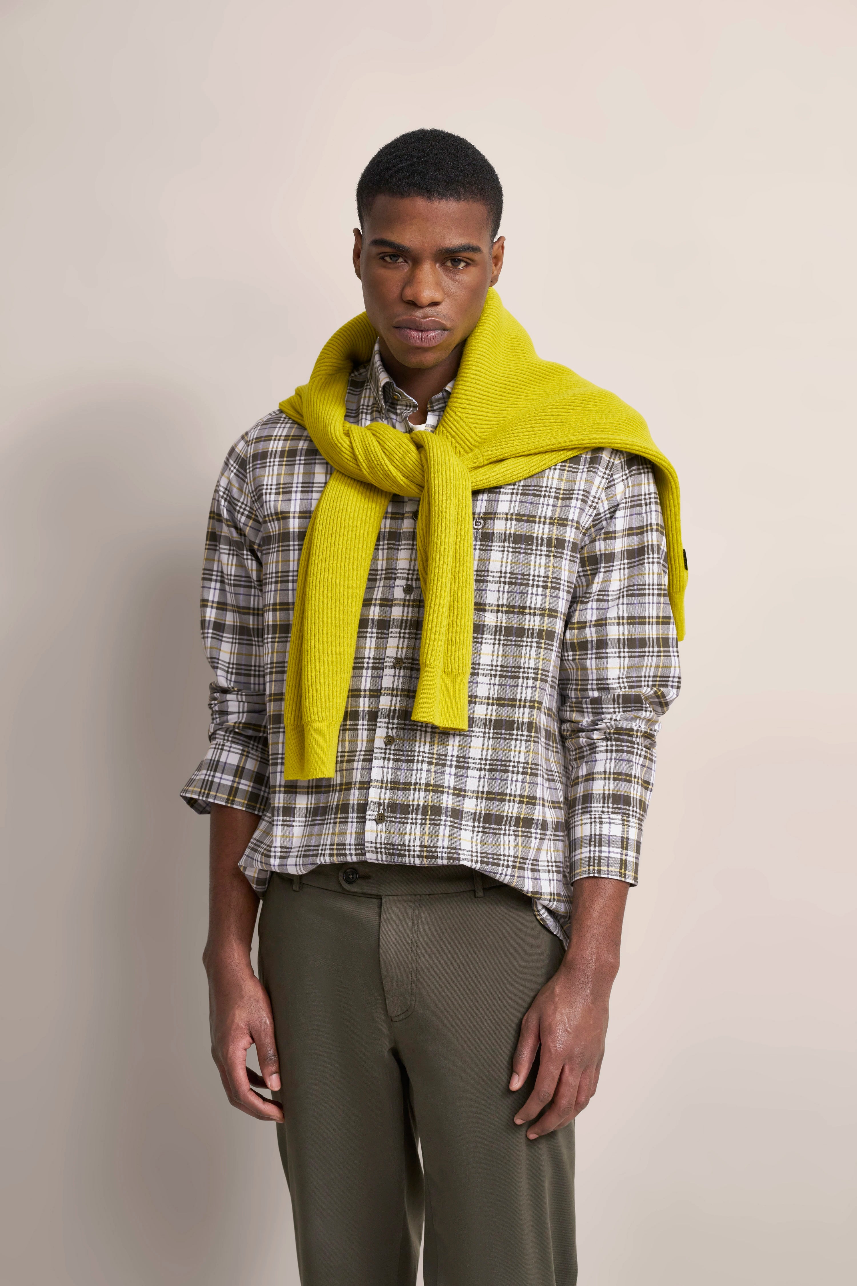 Kariertes Hemd, olivgrüne Hose, gelber Pullover über den Schultern, Modelbild, entspannte Haltung.
