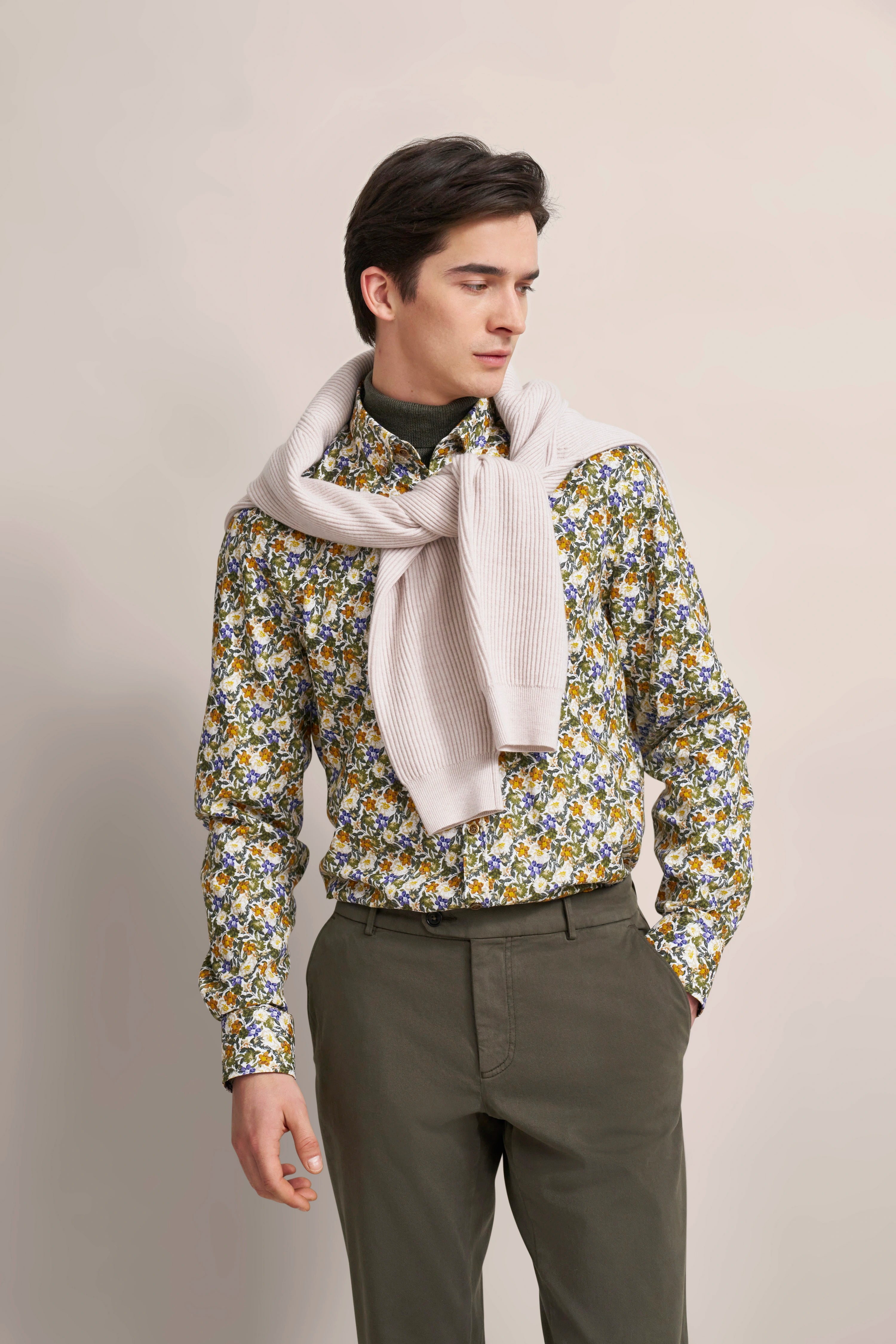 Model trägt florales Hemd, beige Pullover um die Schultern, olivgrüne Hose, seitliches Posing.