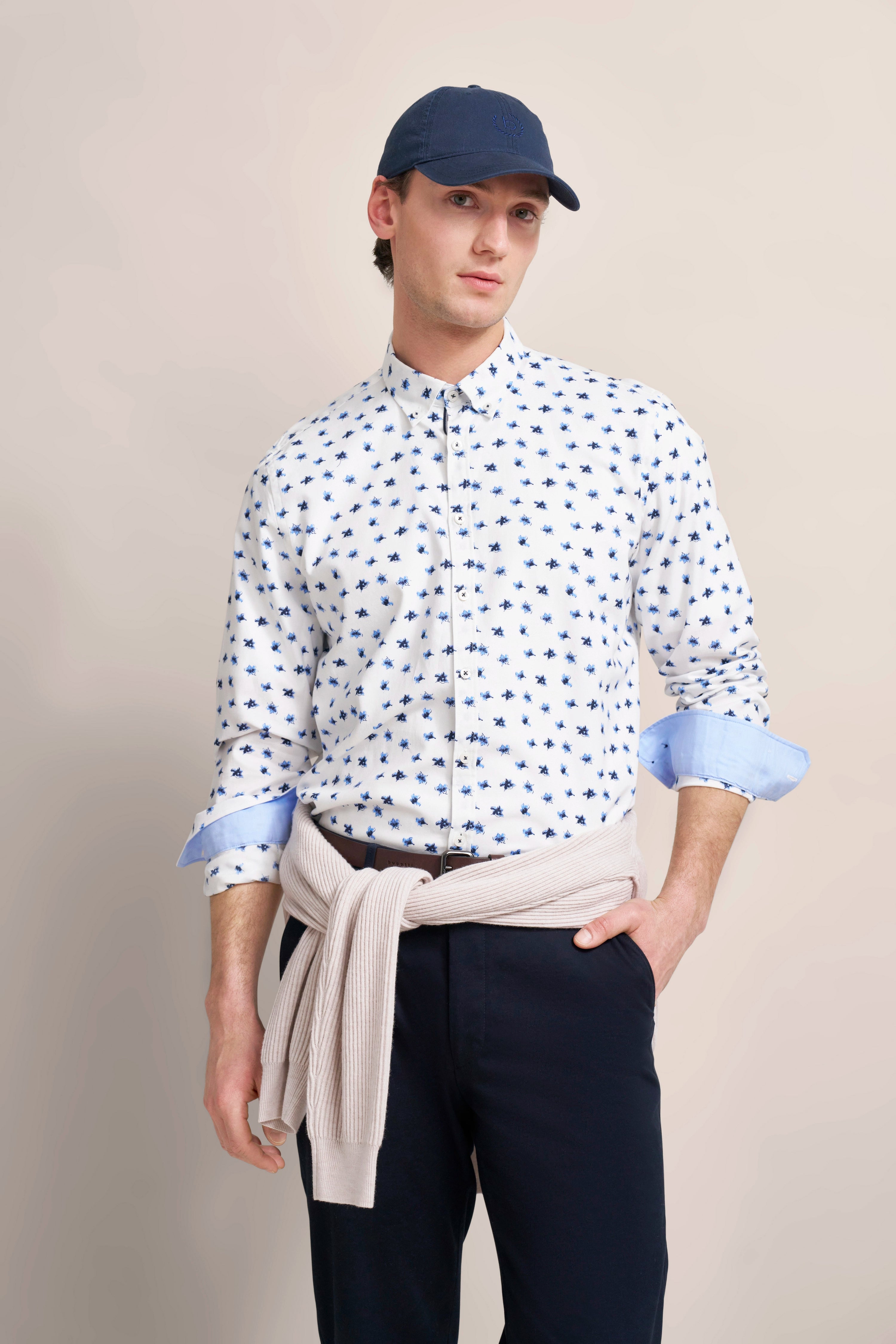 Model in weißem Hemd mit blauem Muster, beiger Pullover um die Taille, dunkelblaue Hose, lässige Pose.