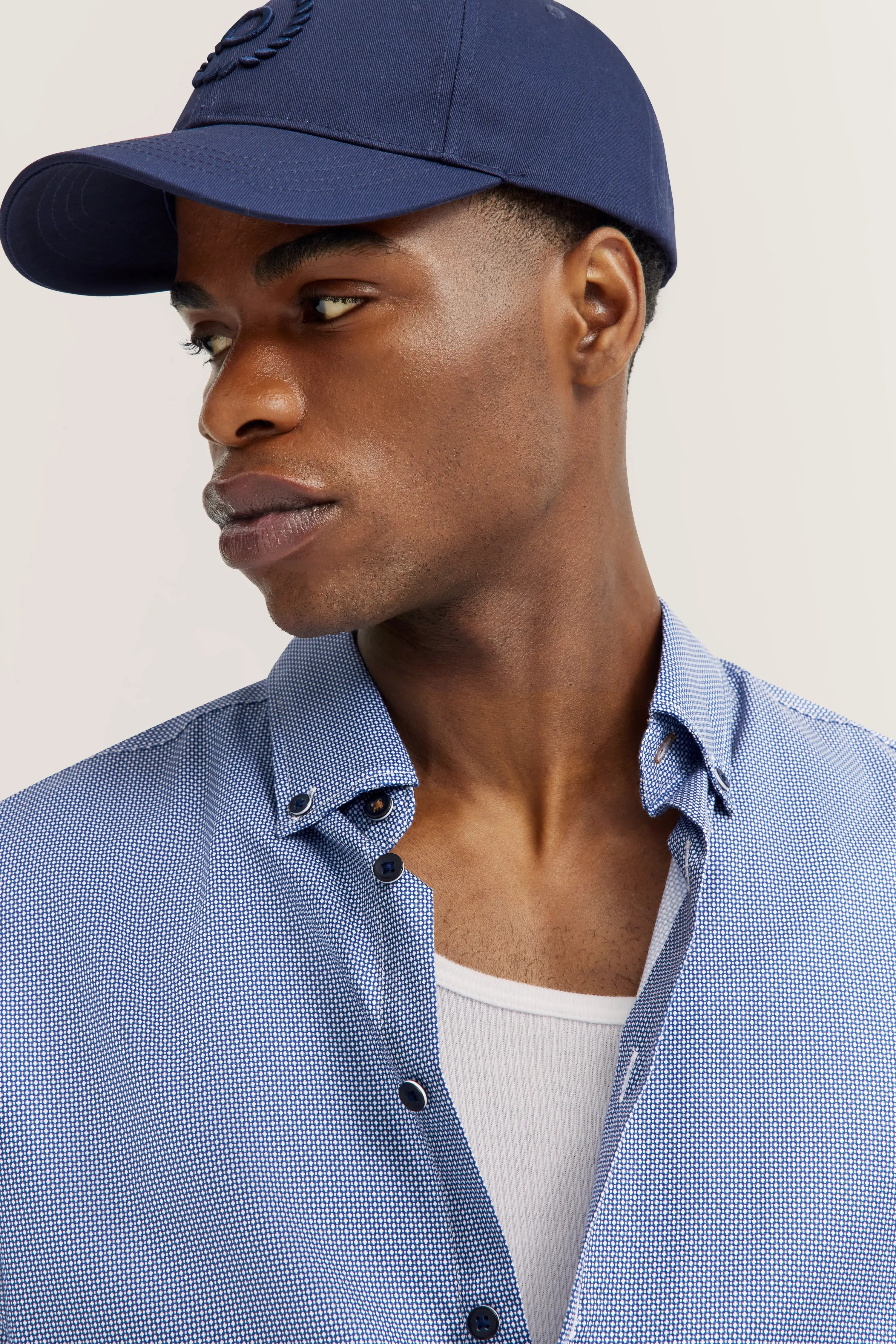 Blauw patroon bugatti overhemd, wit shirt, donkerblauwe pet, model in ontspannen pose.