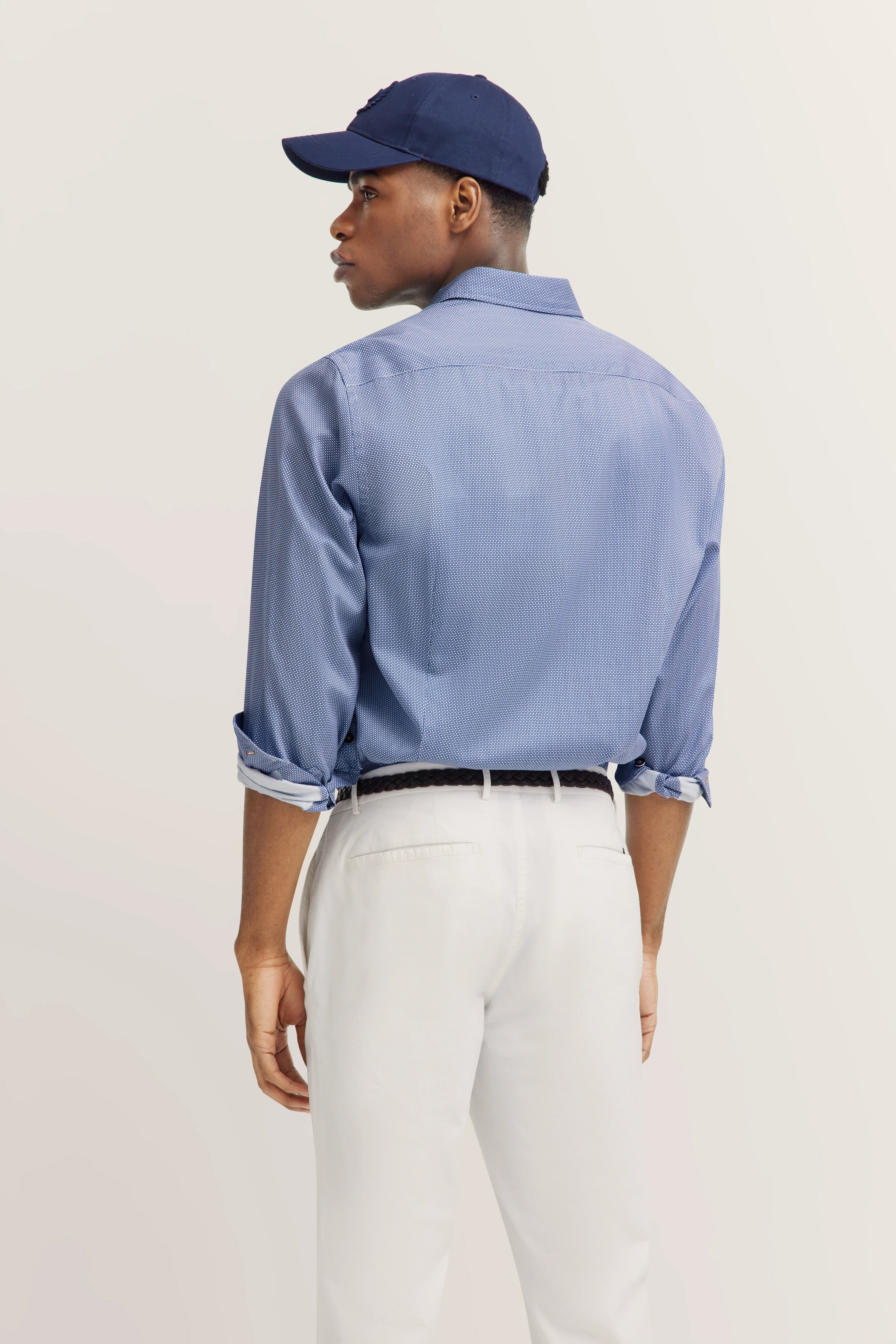Model in blauw shirt met fijn patroon, witte broek, donkerblauwe pet. Achteraanzicht, ontspannen pose.