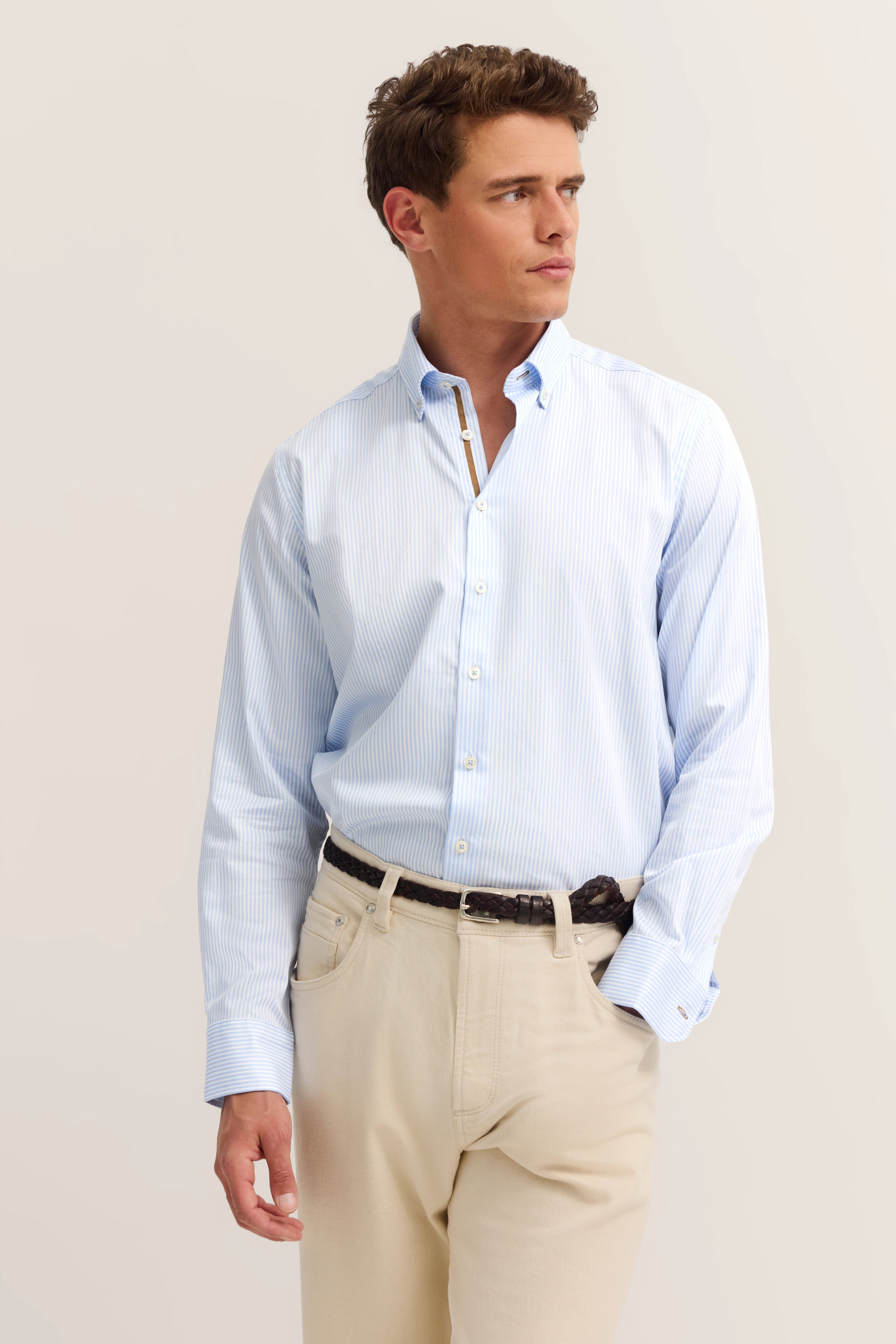 Model in light blue striped shirt and beige trousers. Casual pose, looking to the side.