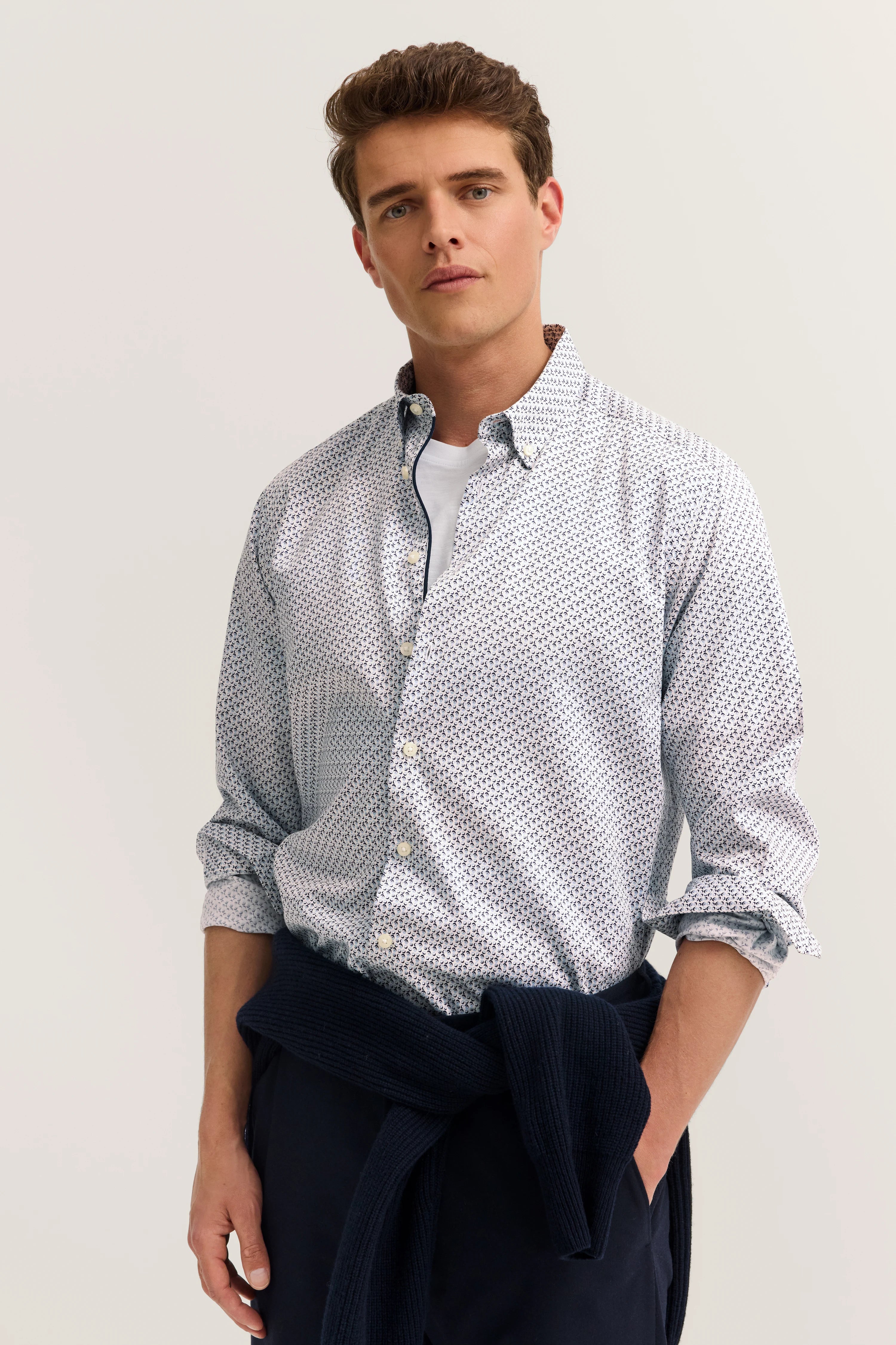 Man in patterned shirt and dark blue trousers, casually with sweater tied around waist.