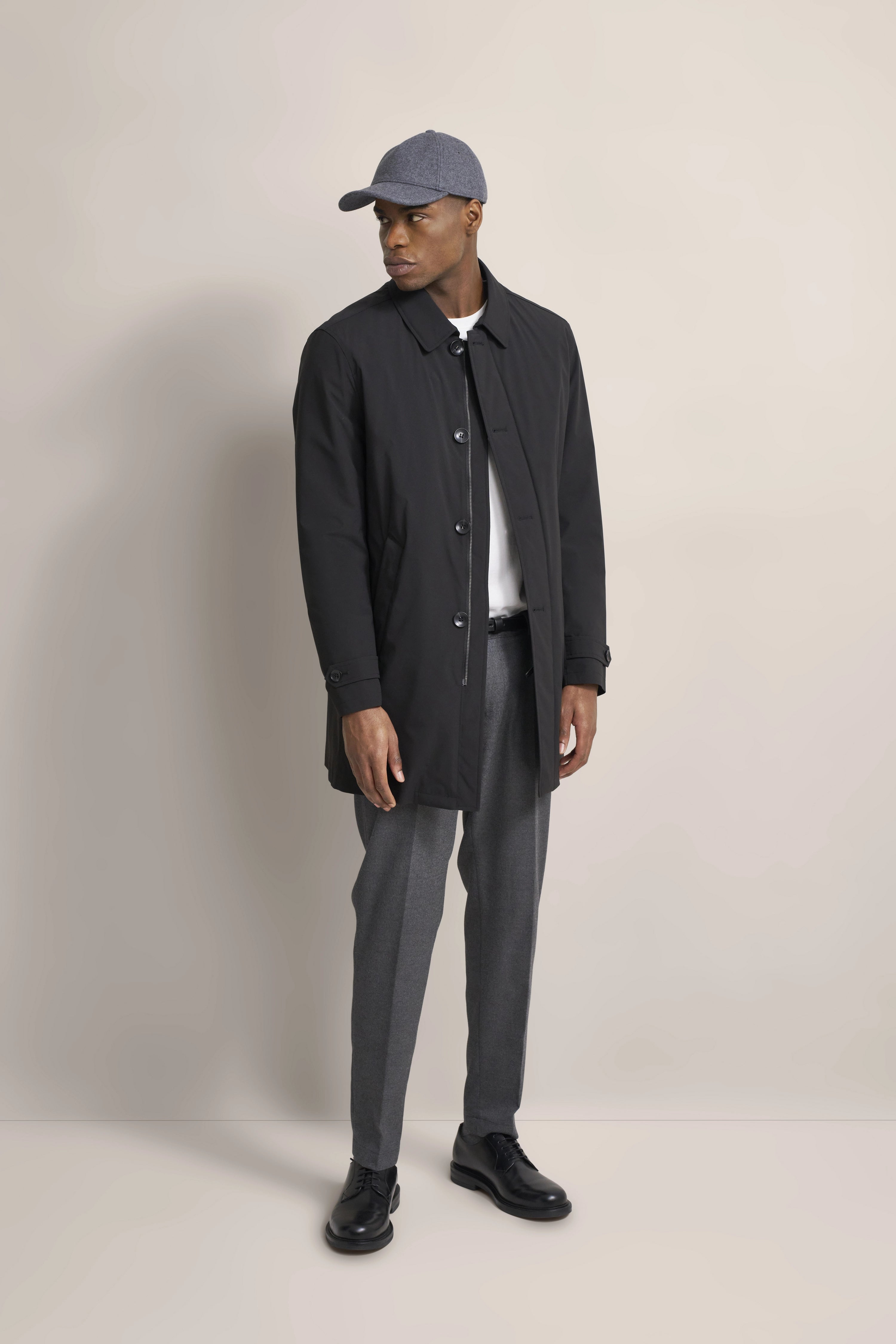 Model trägt schwarzen bugatti Mantel, graue Hose, weiße Oberteil, graue Mütze, schwarze Schuhe in entspannter Pose.