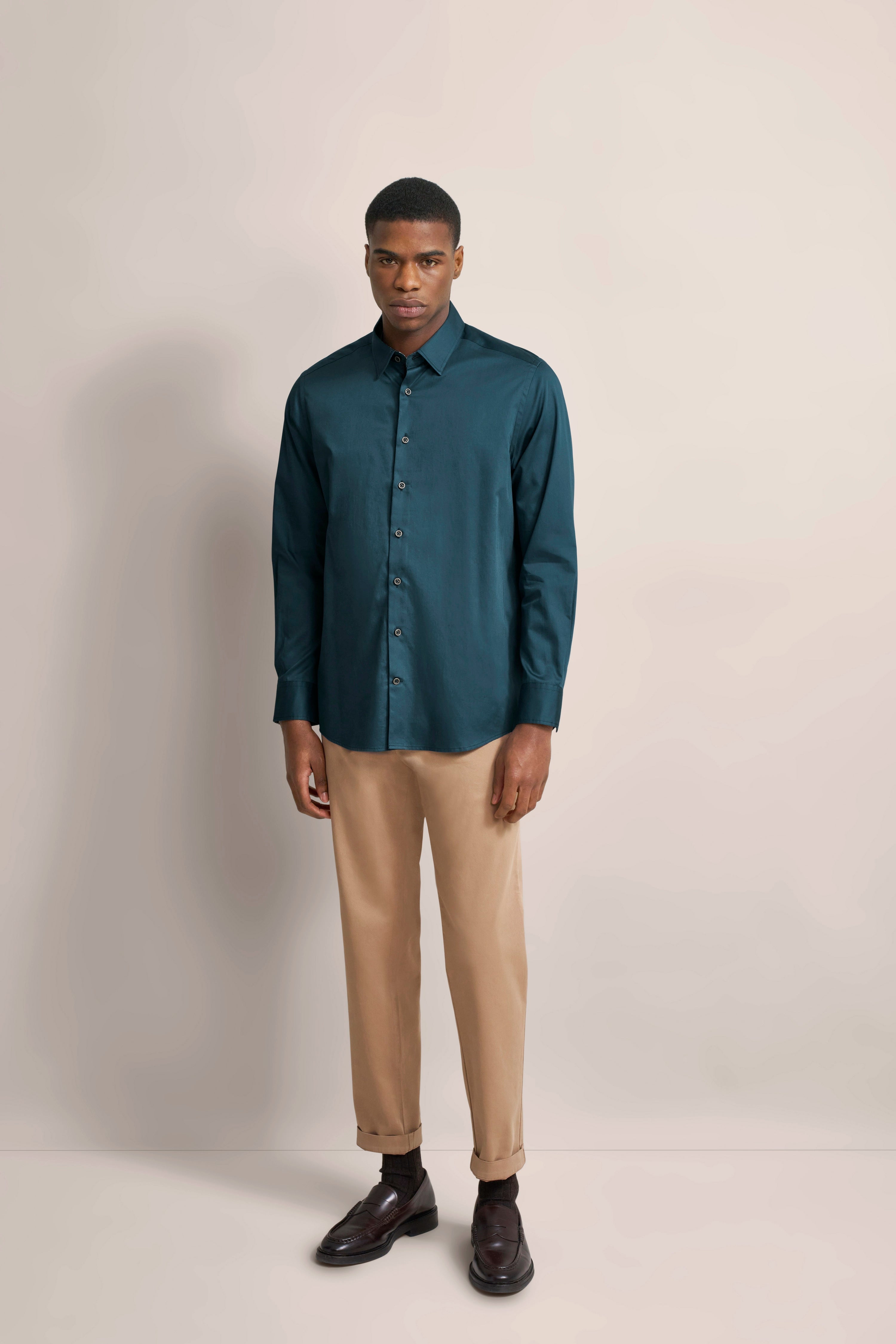 bugatti Modelbild: Mann in petrolfarbenem Hemd und beigen Chinos, entspannt stehend.