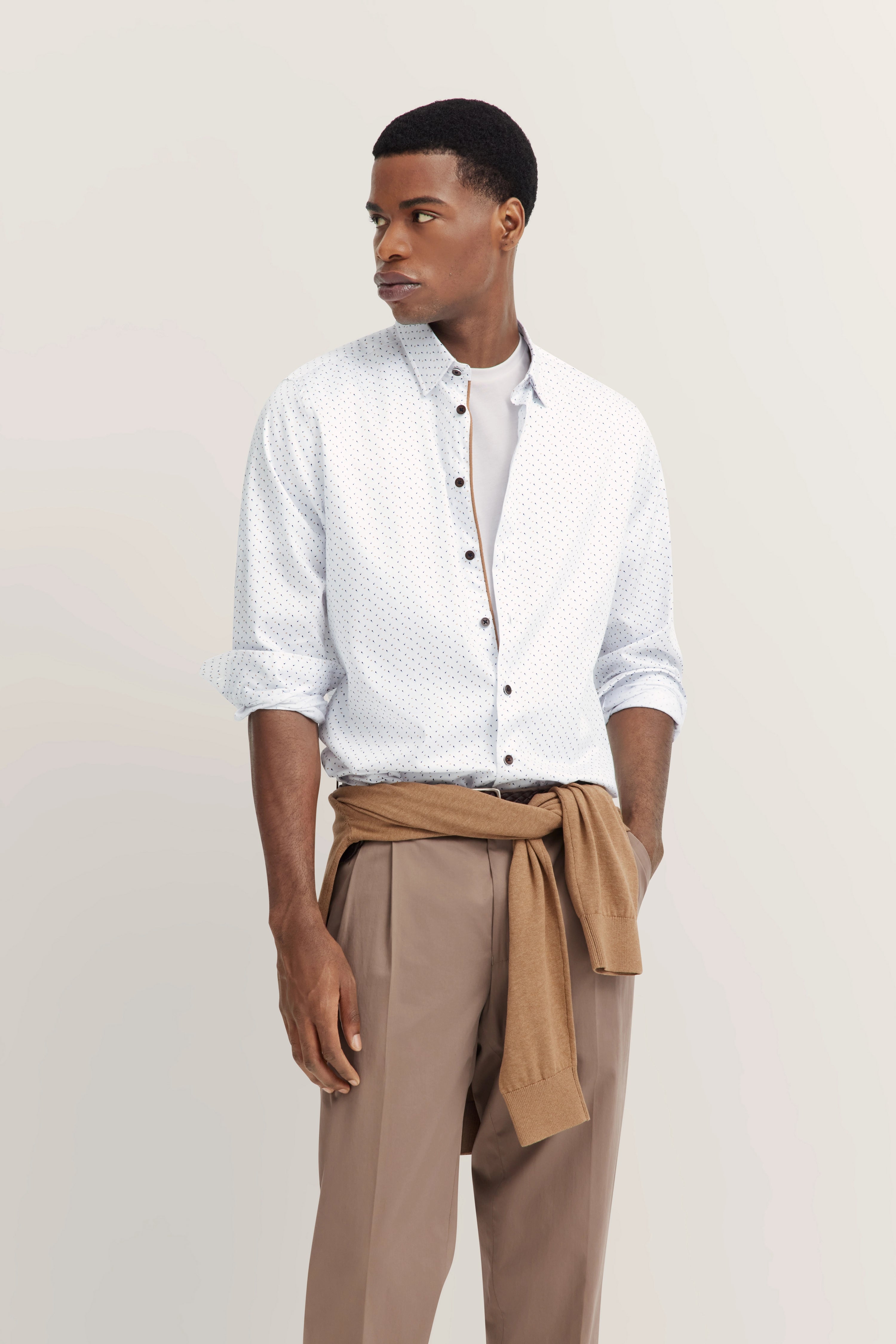 Weißes Punkthemd, hellbraune Hose, beige Pullover um die Hüfte, Model steht entspannt, seitlicher Blick.