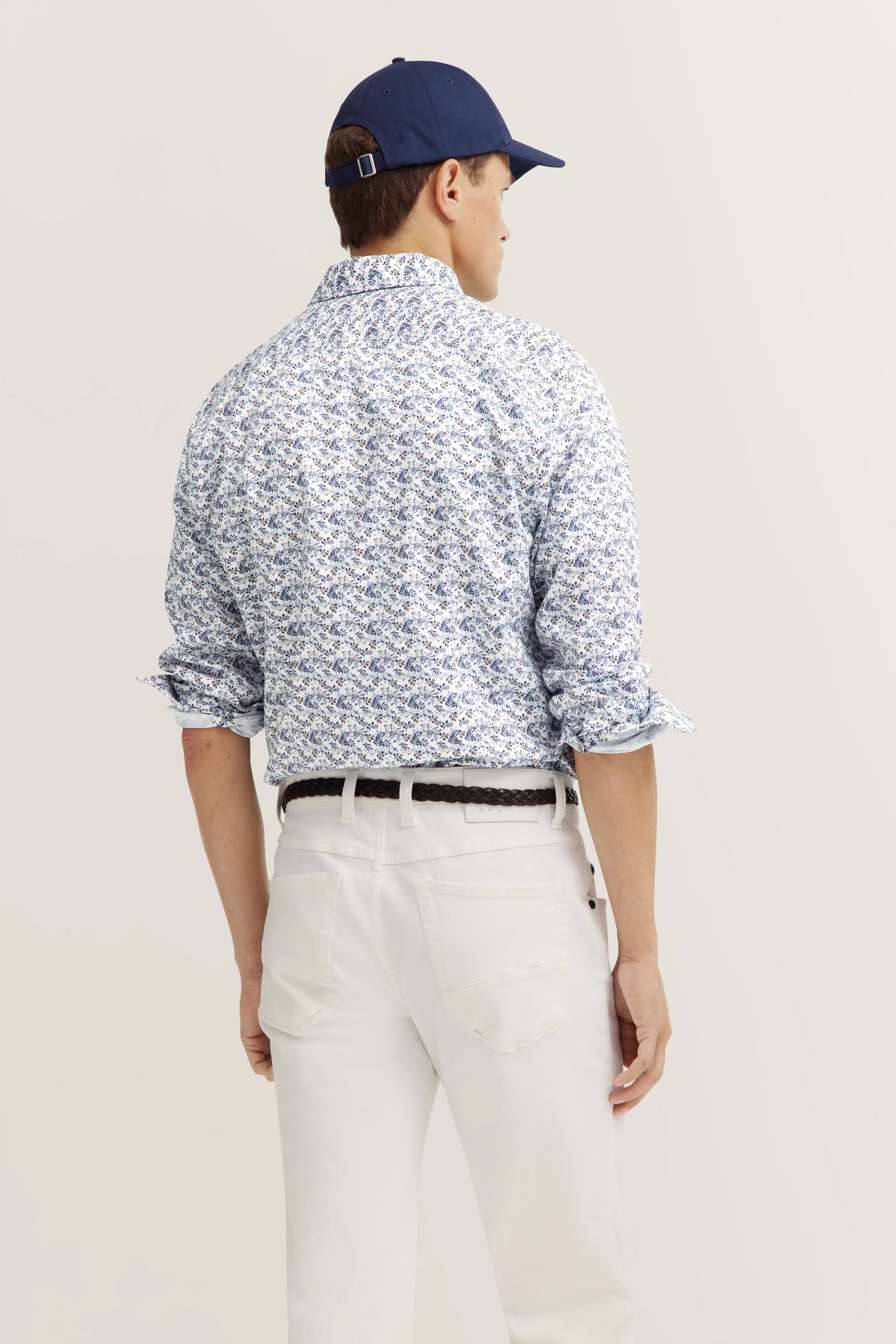 Model in bloemenhemd en witte broek, rugaanzicht, ontspannen pose, blauwe pet.