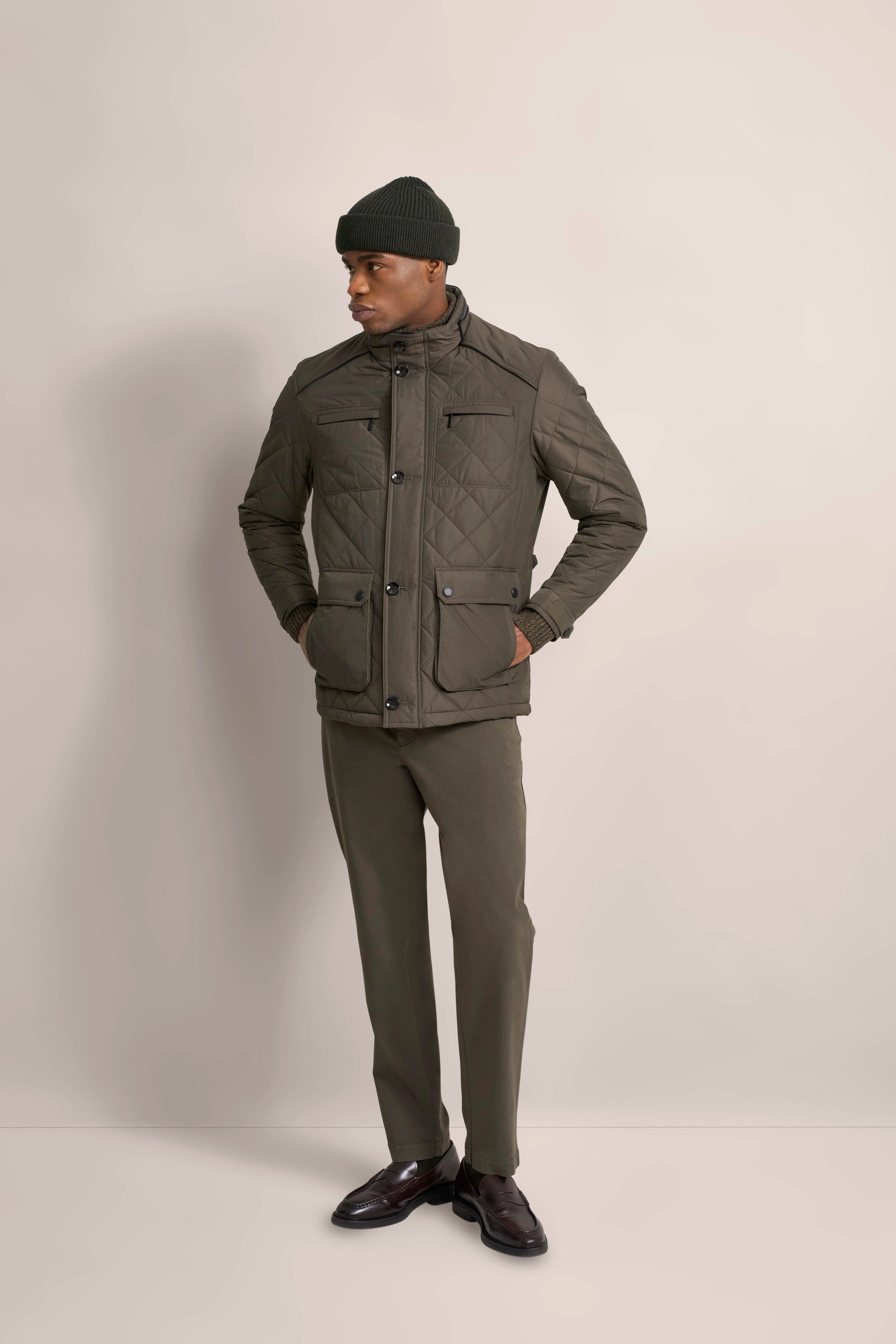 Model in olivgrüner Steppjacke, passender Hose, Mütze und braunen Schuhen, Hände in Taschen.