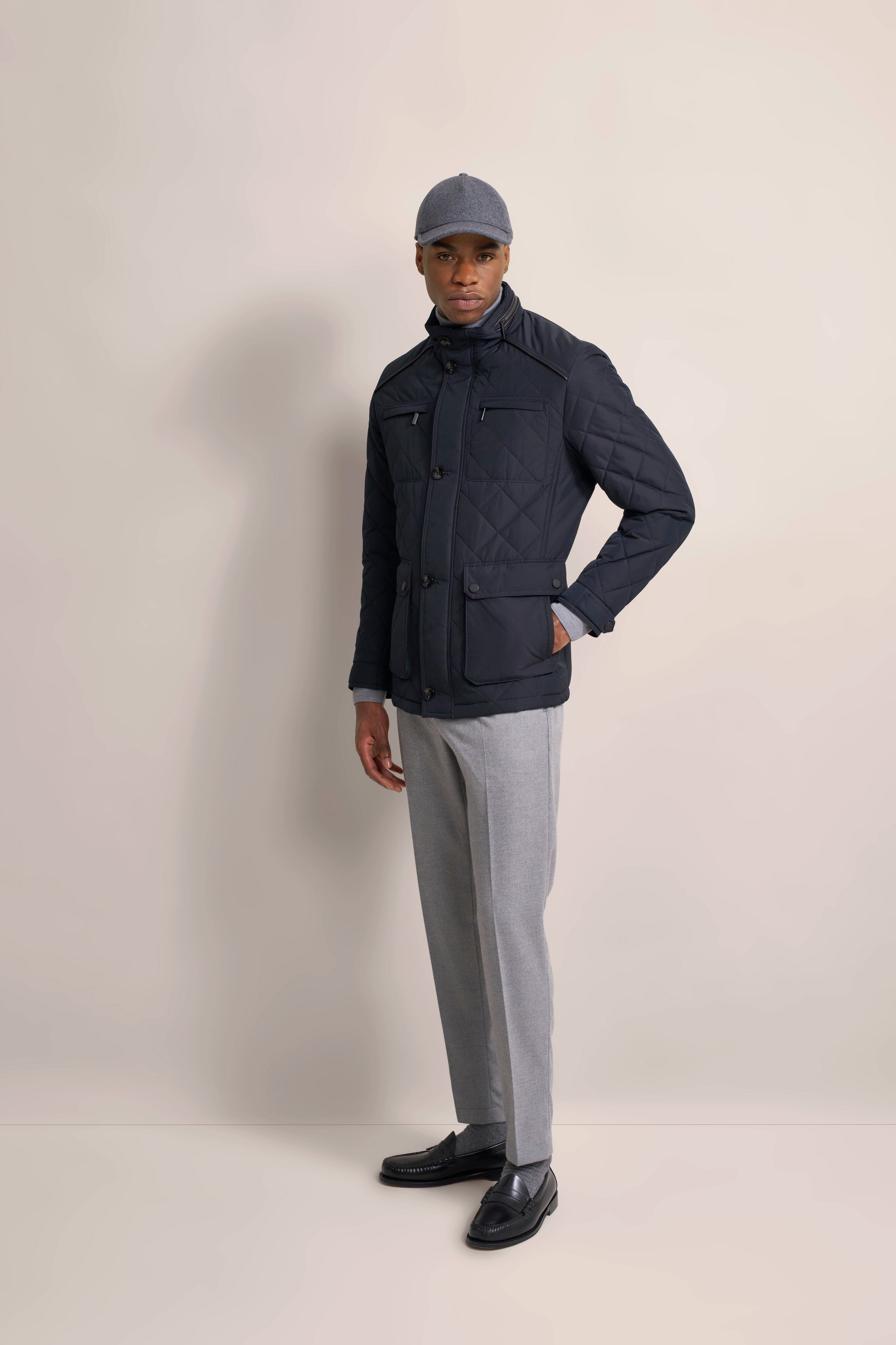Model in dunkelblauer Steppjacke, grauer Hose, grauer Mütze, legeres Posing.