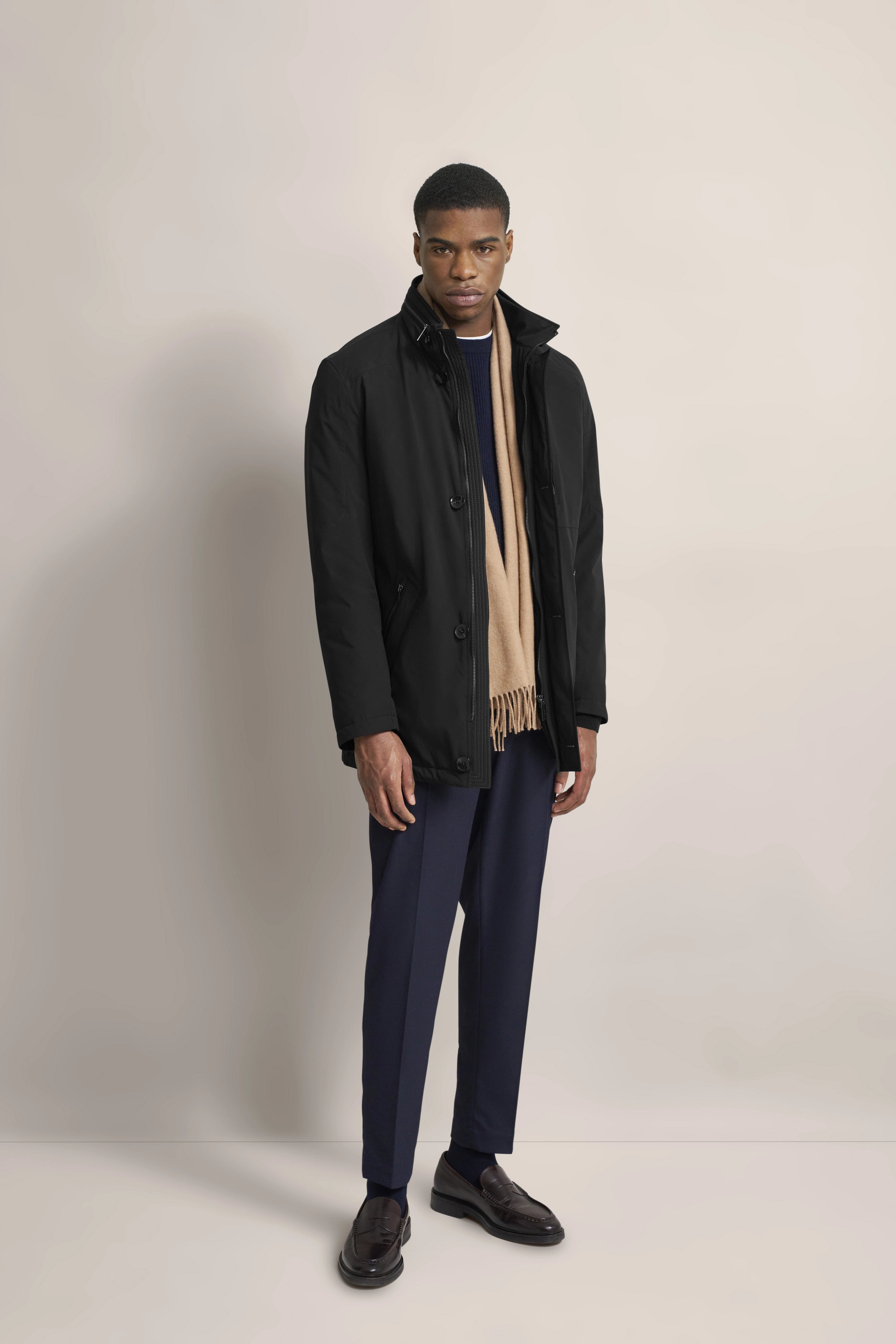 Model in schwarzer Jacke, blauer Hose, beigem Schal, elegant posierend. bugatti Stil und Qualität.