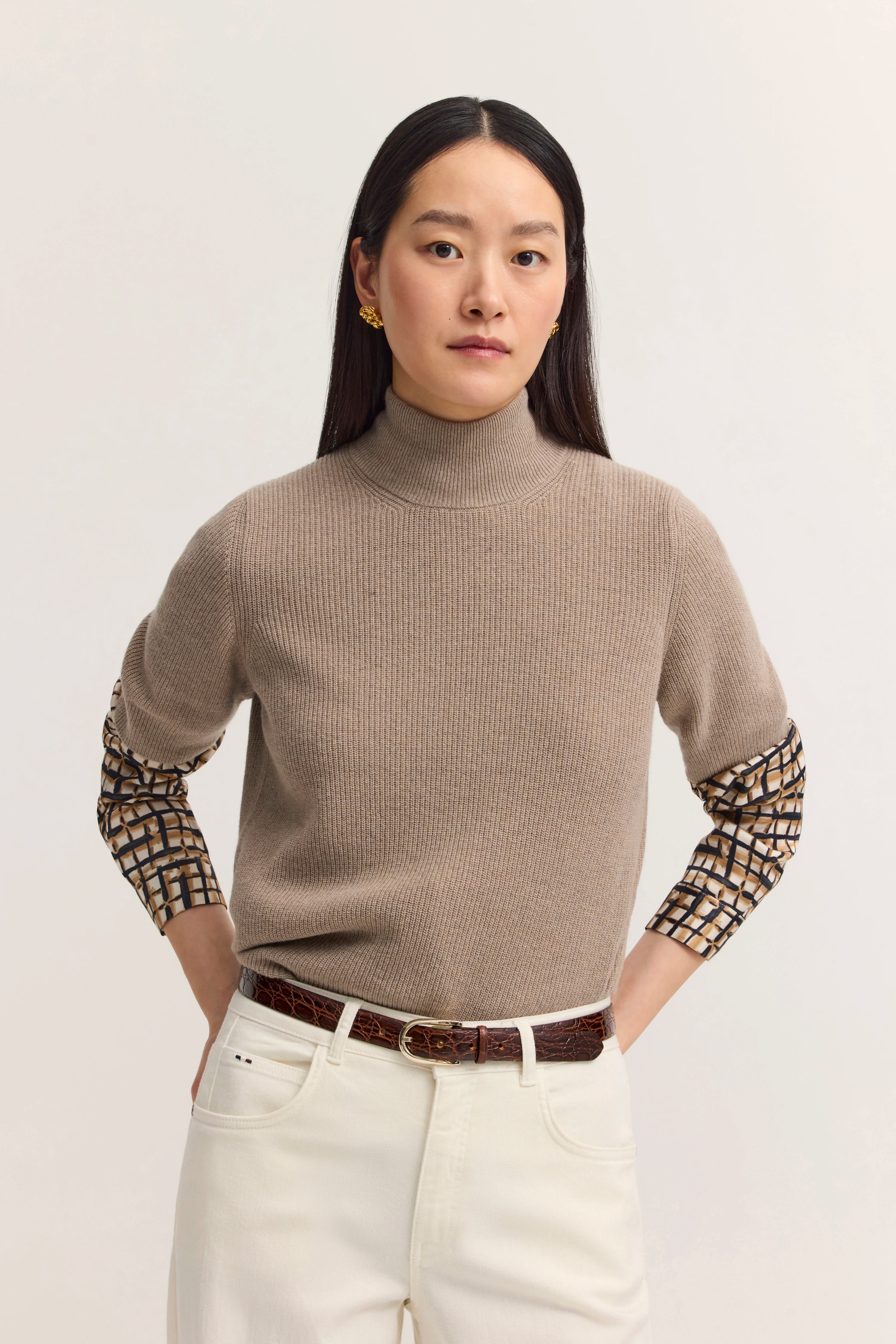 Modelbild: Frau in beige Rollkragenpullover, gemusterte Ärmel, weiße Hose. Hände in Taschen, entspannte Haltung.