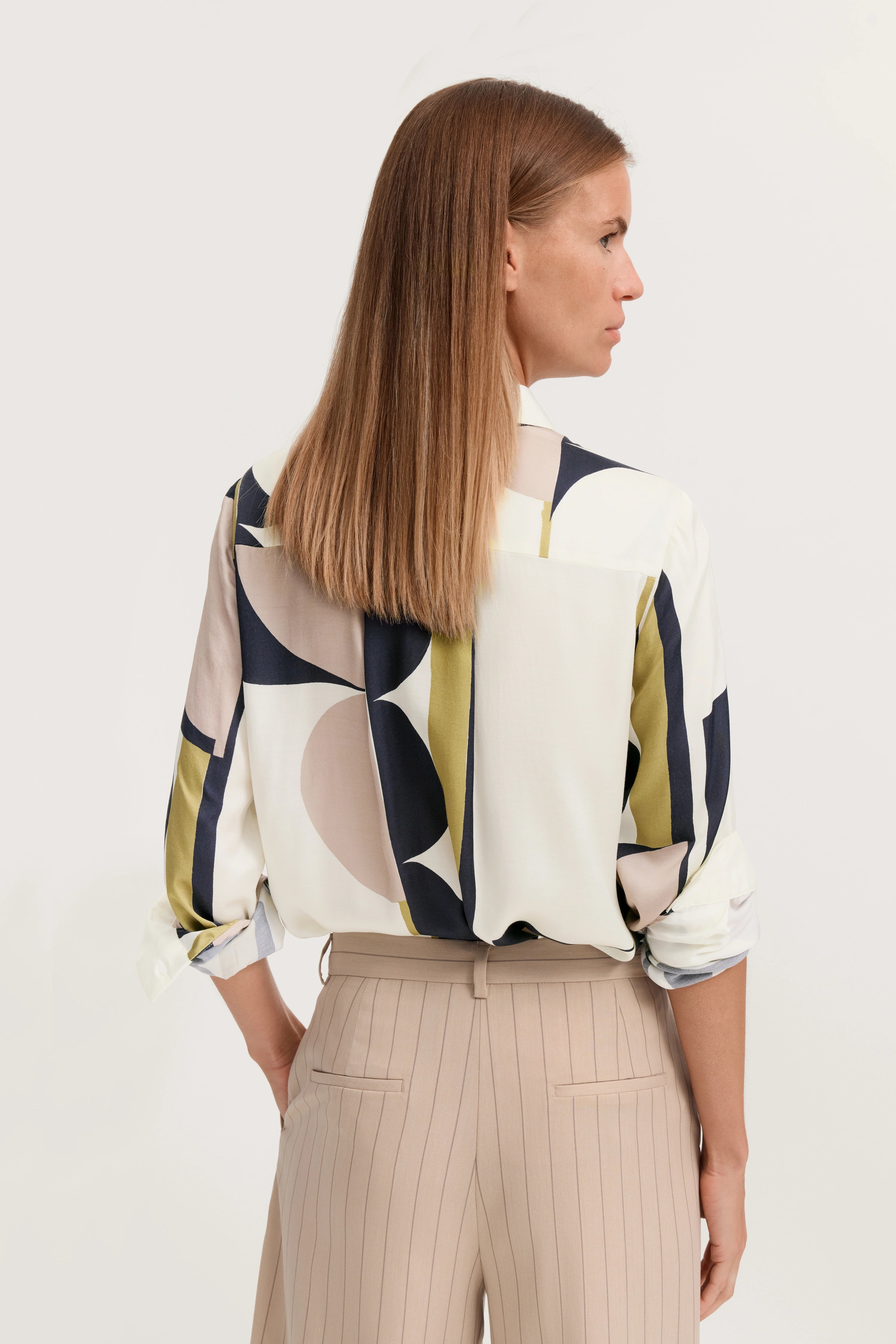 Bluse mit geometrischem Muster in Beige, Weiß, Blau, Oliv. Beige gestreifte Hose, Model von hinten.
