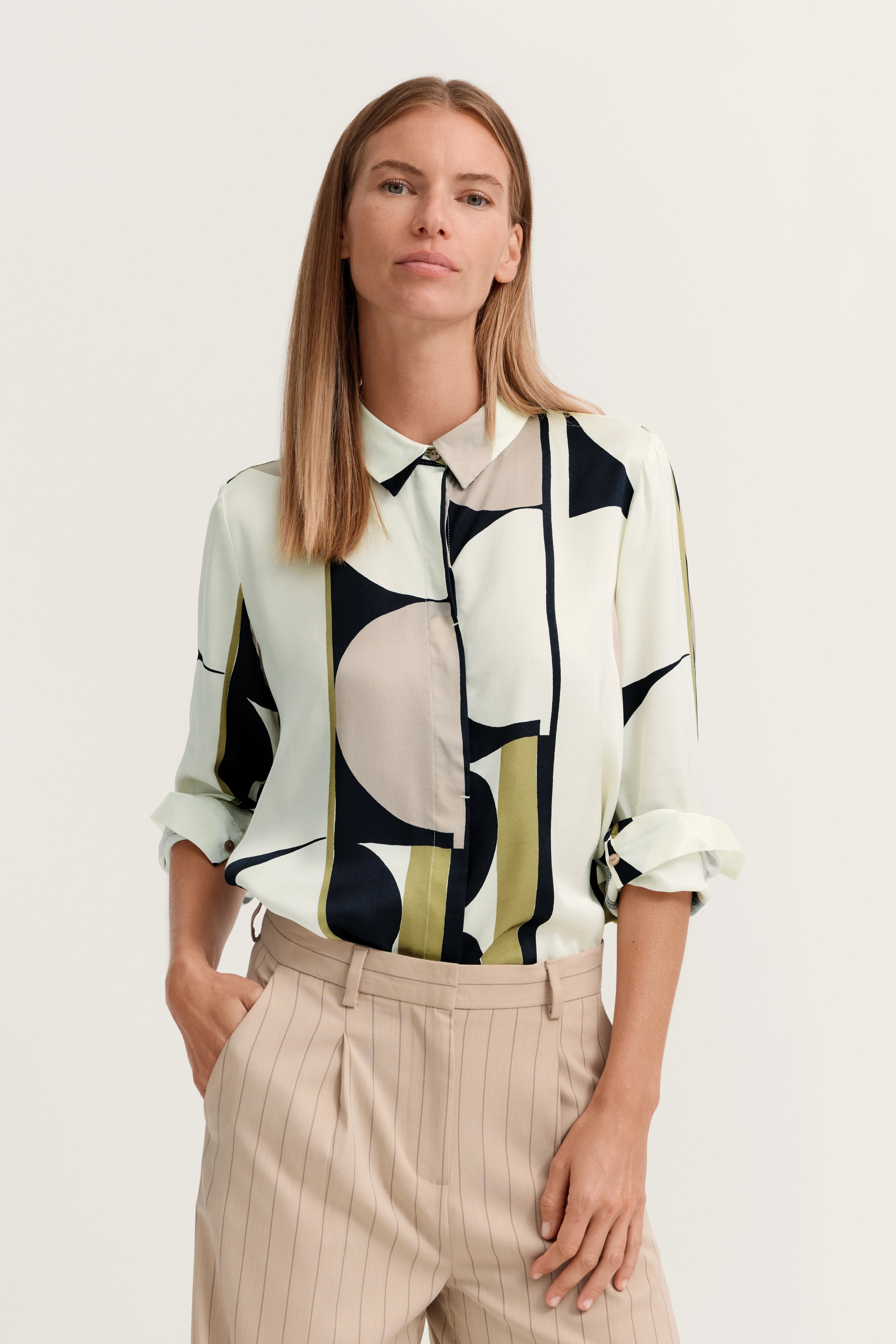 Elegante Bluse mit geometrischen Mustern, Modelbild stehend, Hände in Hosentaschen, entspannter Look.