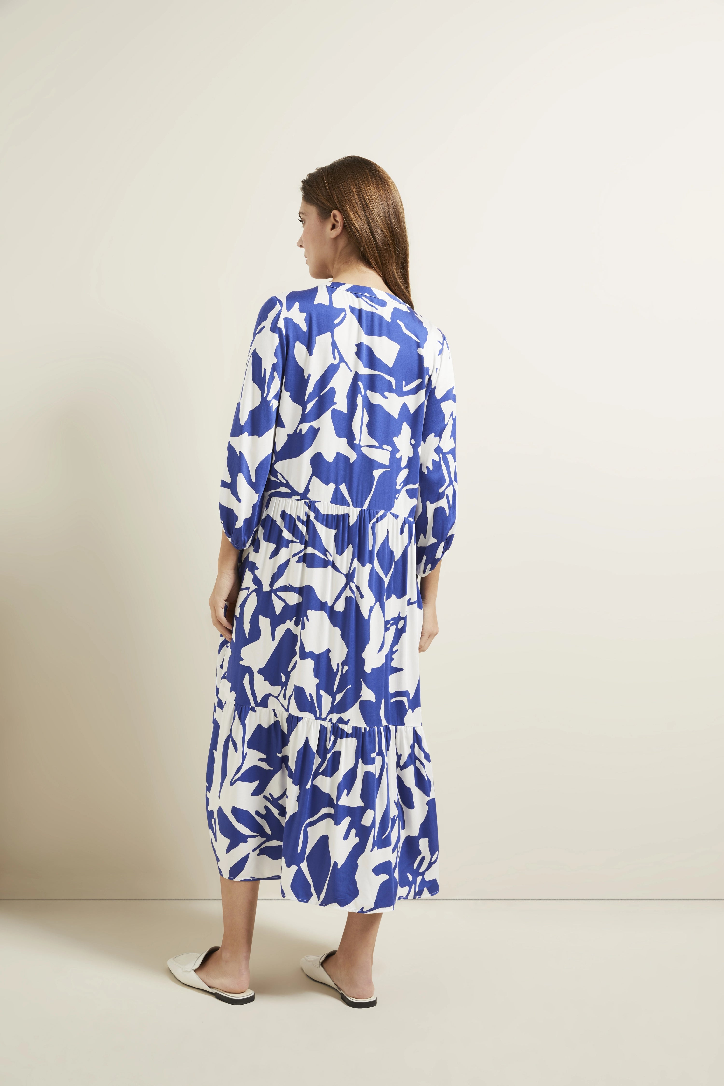 Model in blau-weiß gemustertem bugatti-Kleid, Rückenansicht, lässige Pose, weiße Slipper.