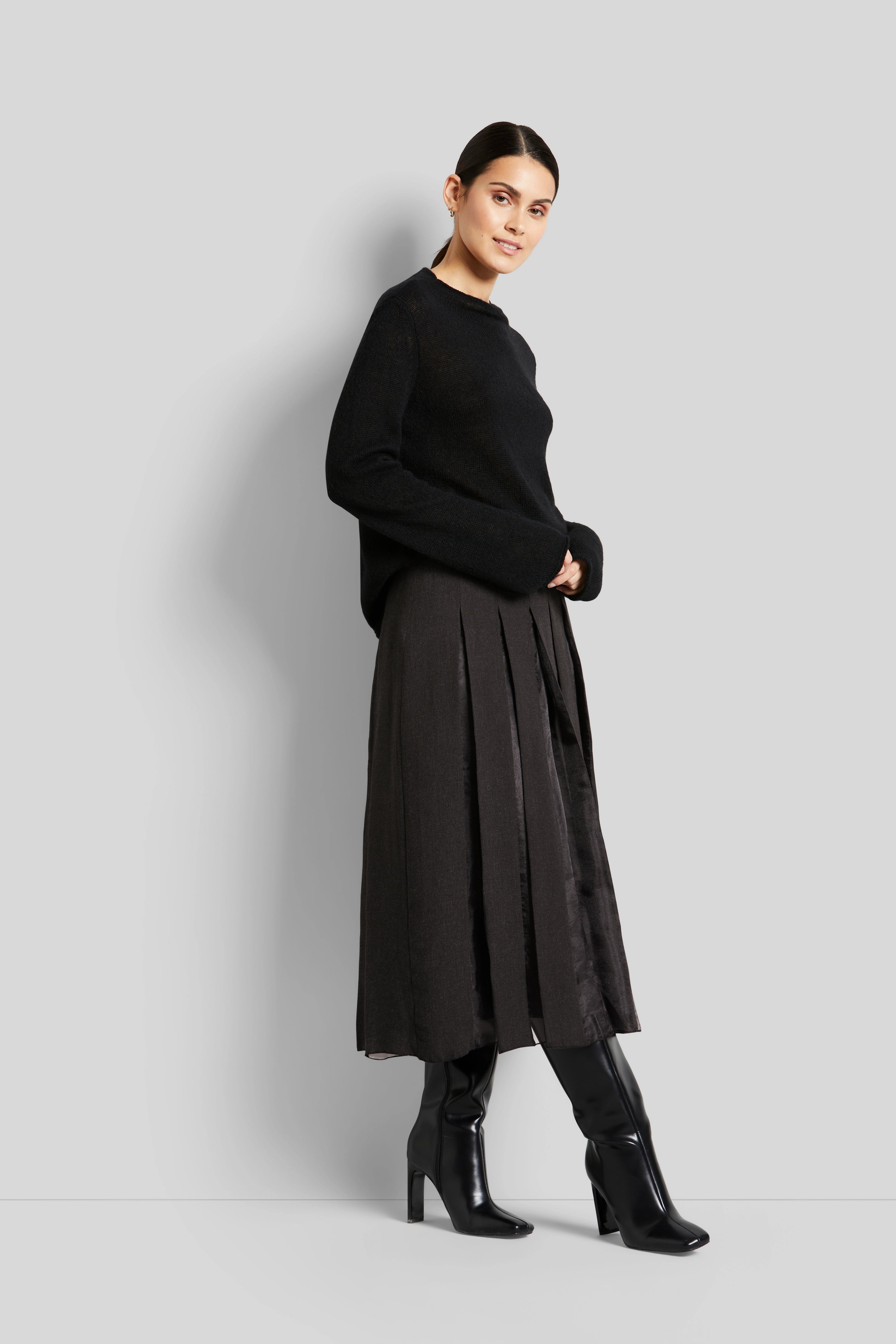Schwarzer bugatti Pullover mit grauem Faltenrock, Modelbild, seitliche Pose, schwarze Stiefel.