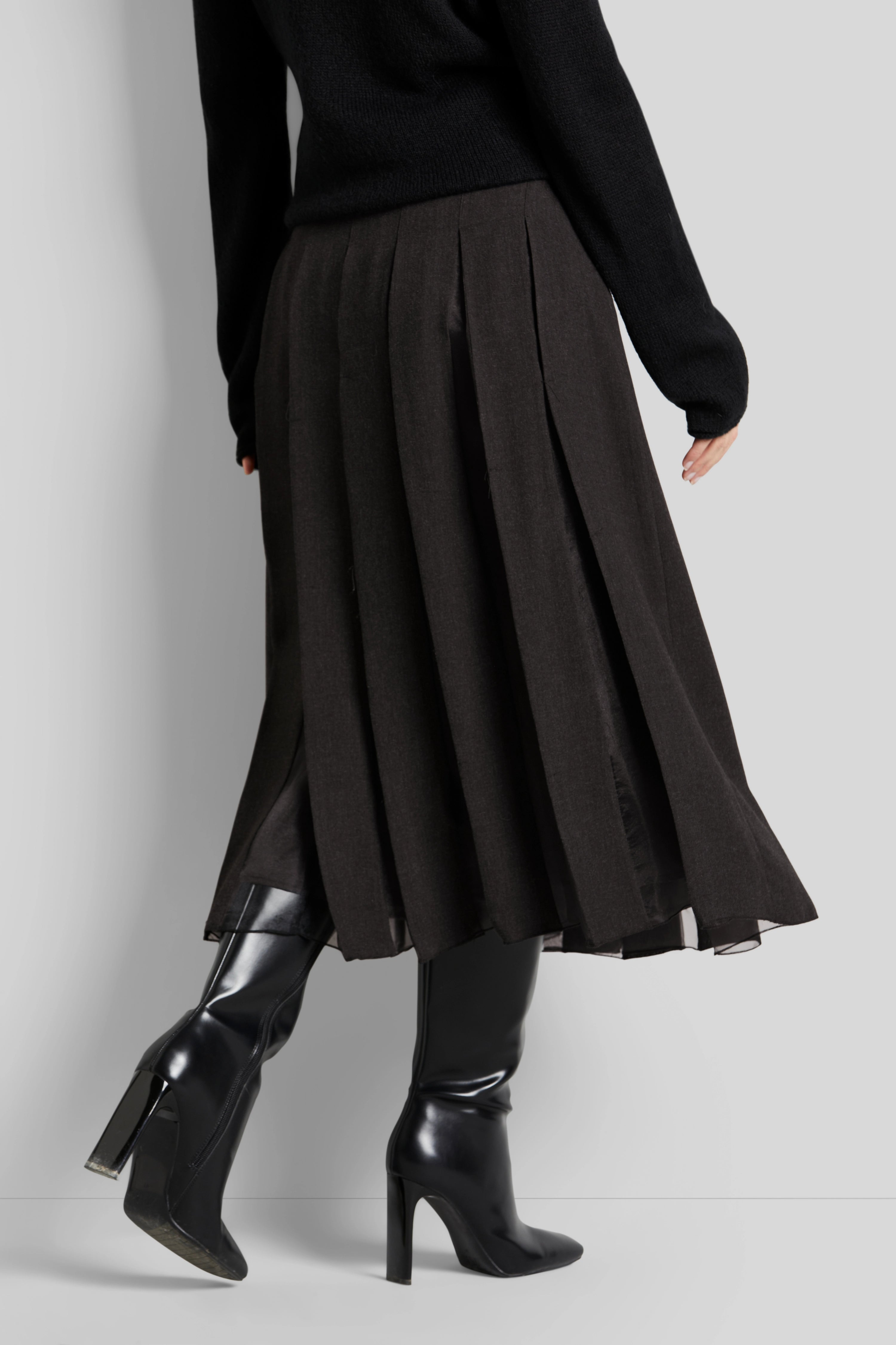 Schwarzer Pullover mit braunem Faltenrock und schwarzen Stiefeln. Model seitlich in Bewegung.