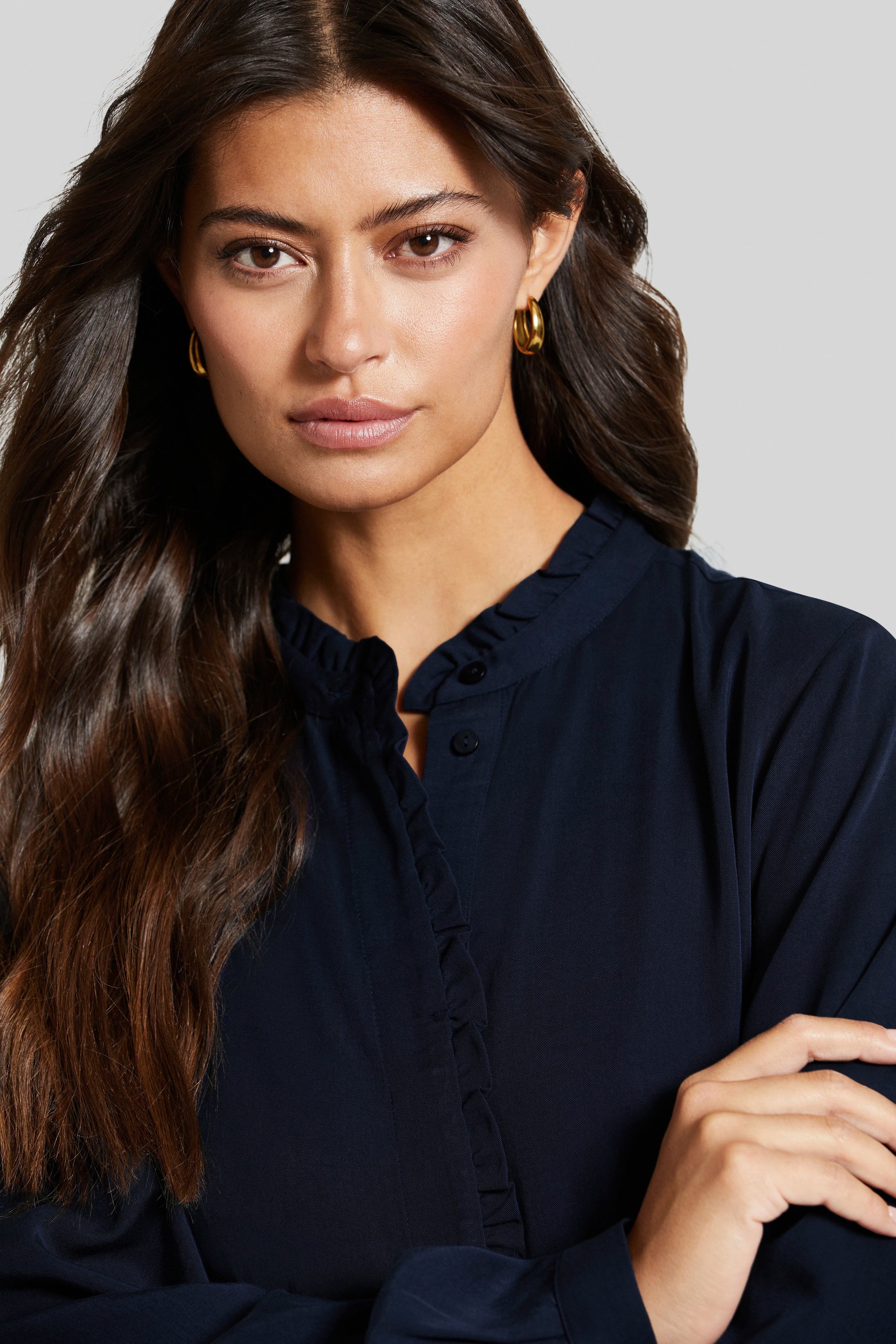 Bust of a woman in a dark blue bugatti blouse, wearing gold earrings.