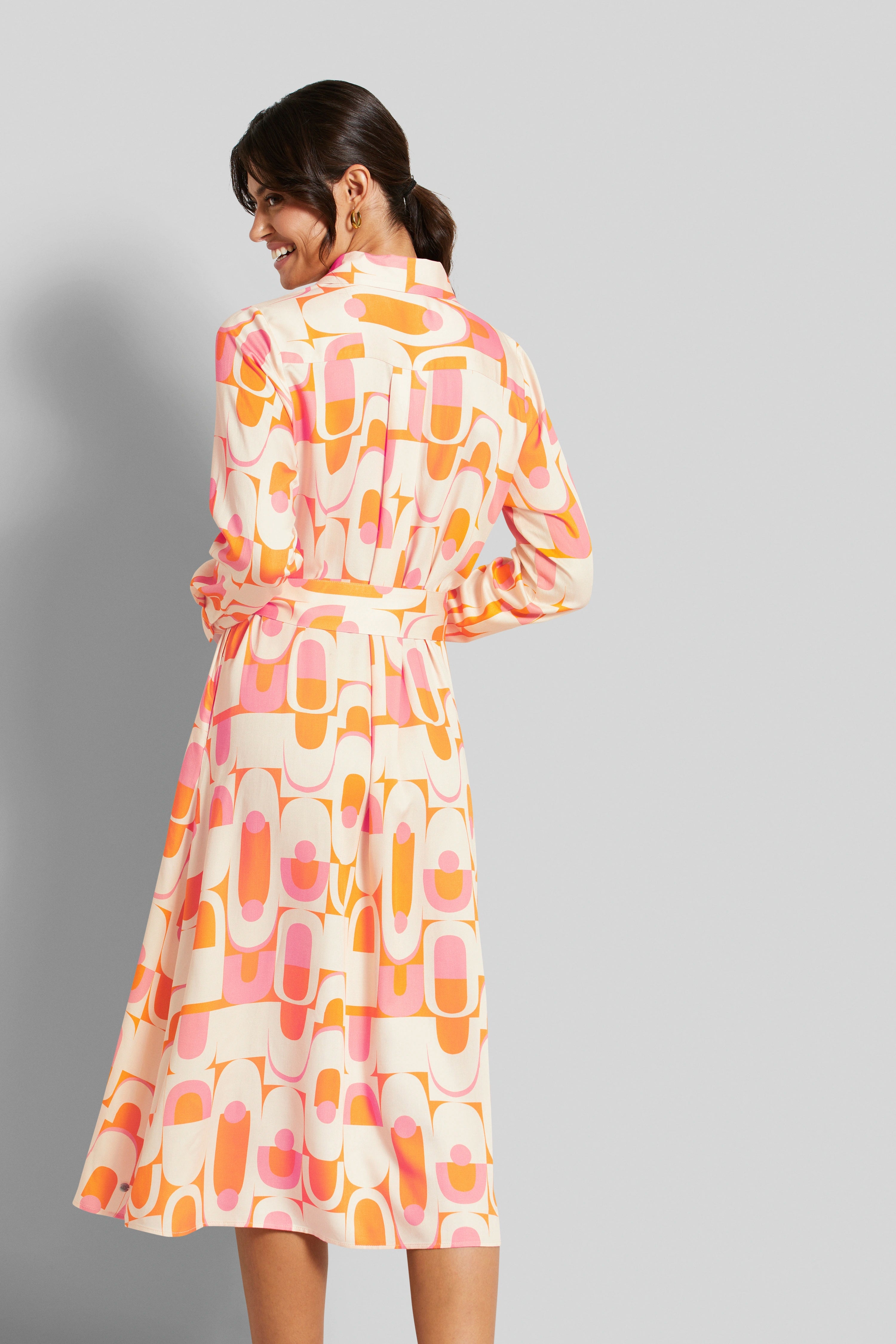 Woman in patterned orange and pink dress, smiling over shoulder. Model image.