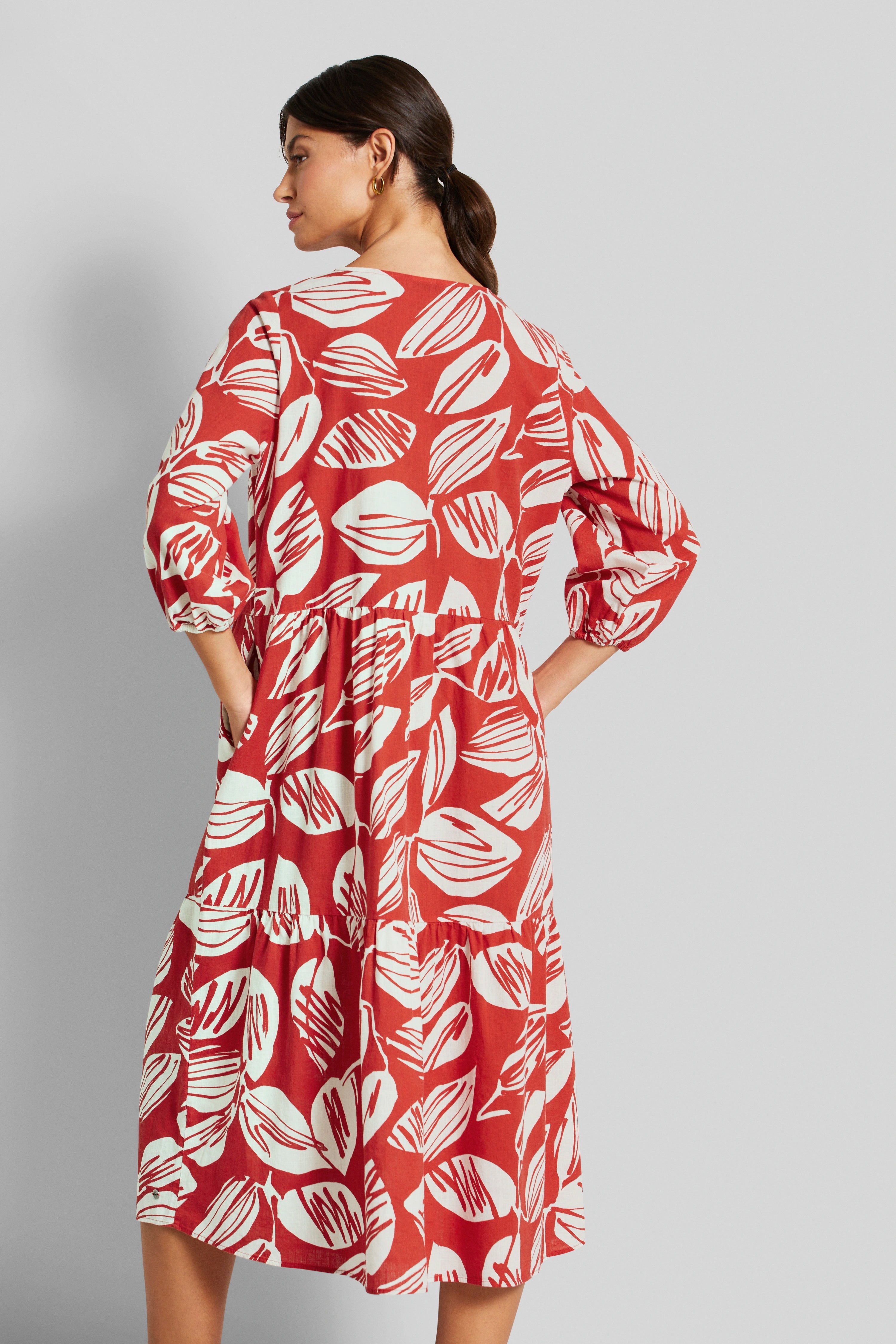 Rotes Kleid mit Blattmuster, Model seitlich stehend, Hände in Taschen, lässige Pose.