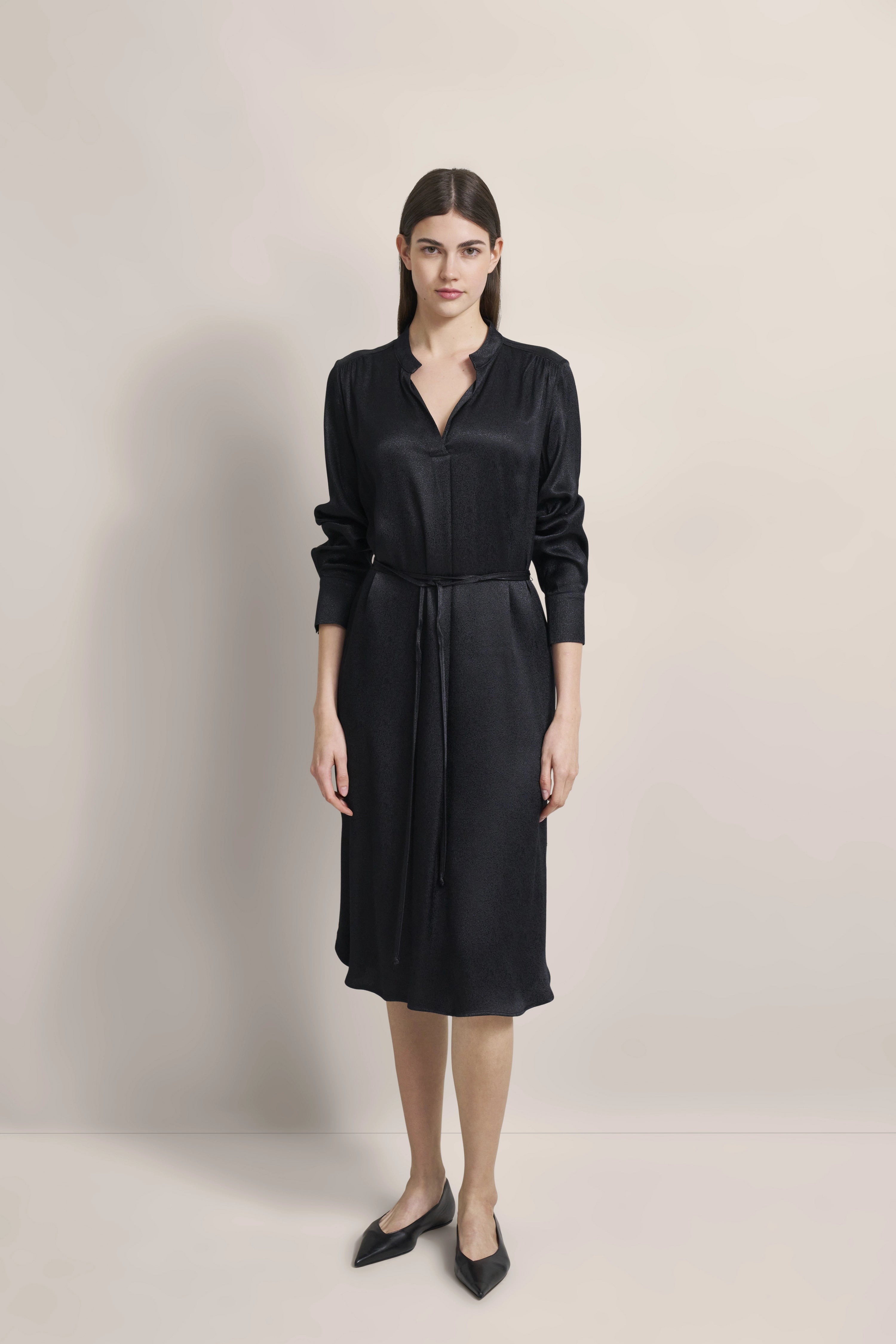 Model in black bugatti dress, long sleeves, waist belt. Elegant pose, standing straight.