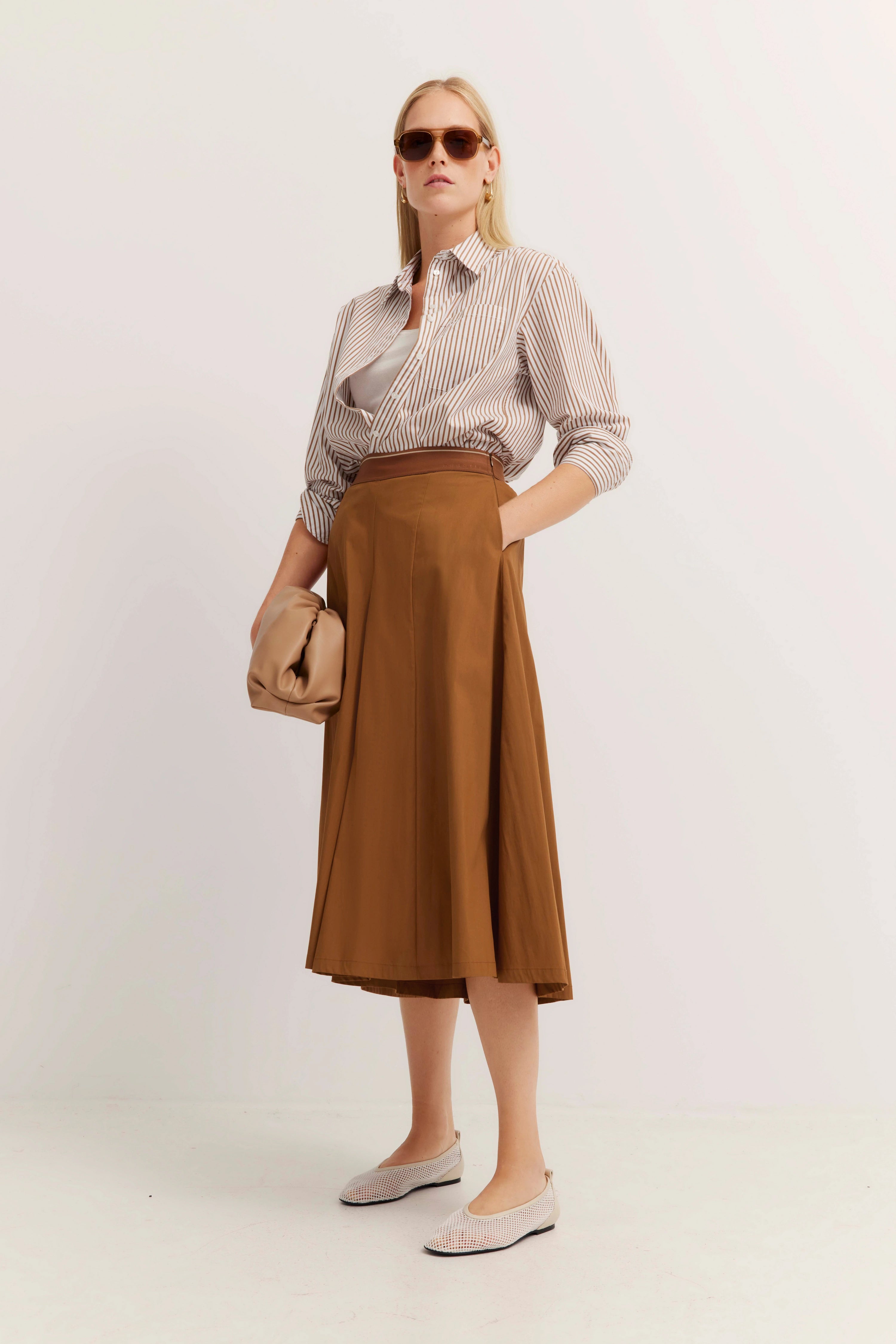 Model draagt gestreepte blouse, bruine midi-rok, beige accessoires, ontspannen houding.