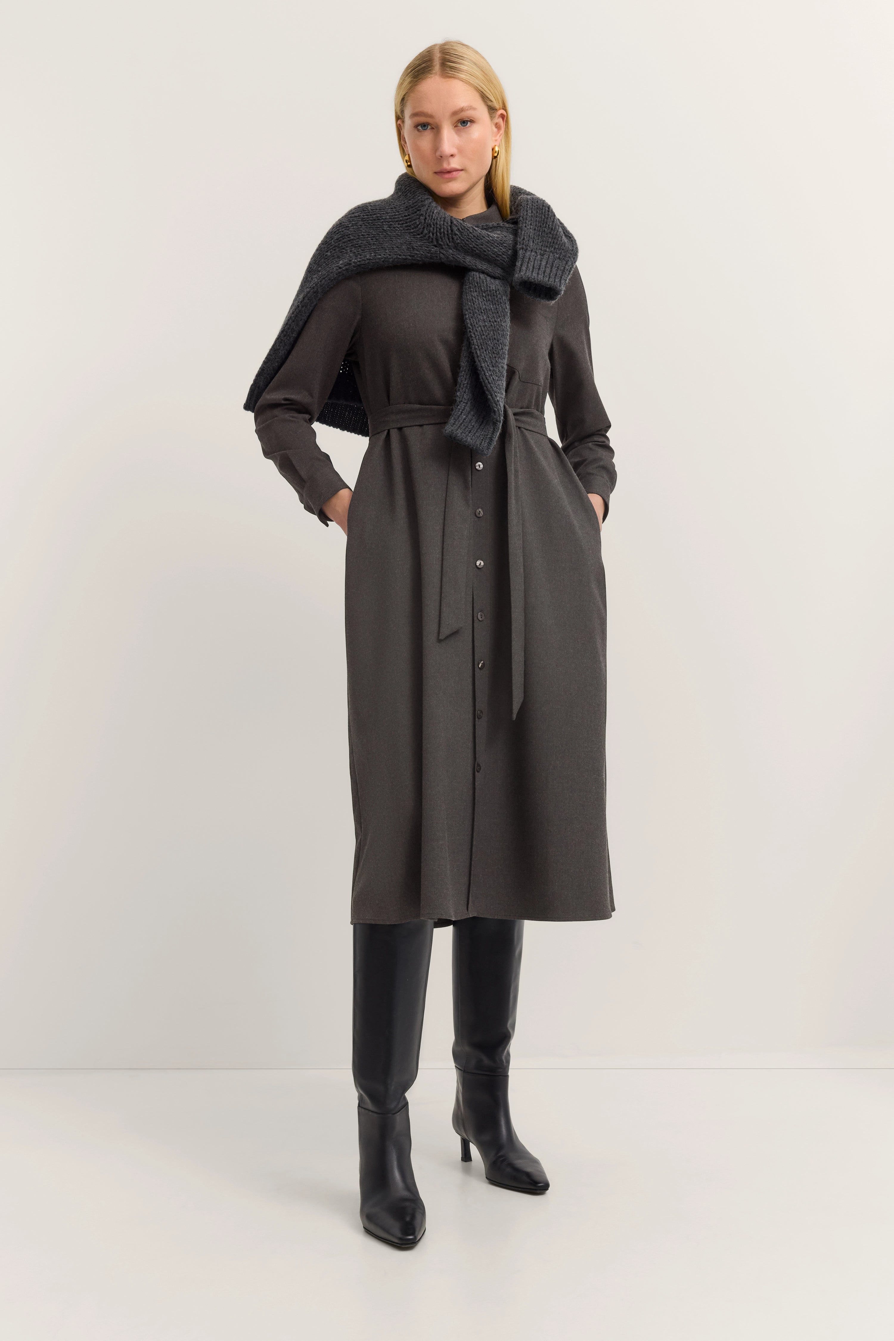 Graues Hemdblusenkleid mit Gürtel, dunkler Schal, schwarze Stiefel, Model posiert aufrecht.