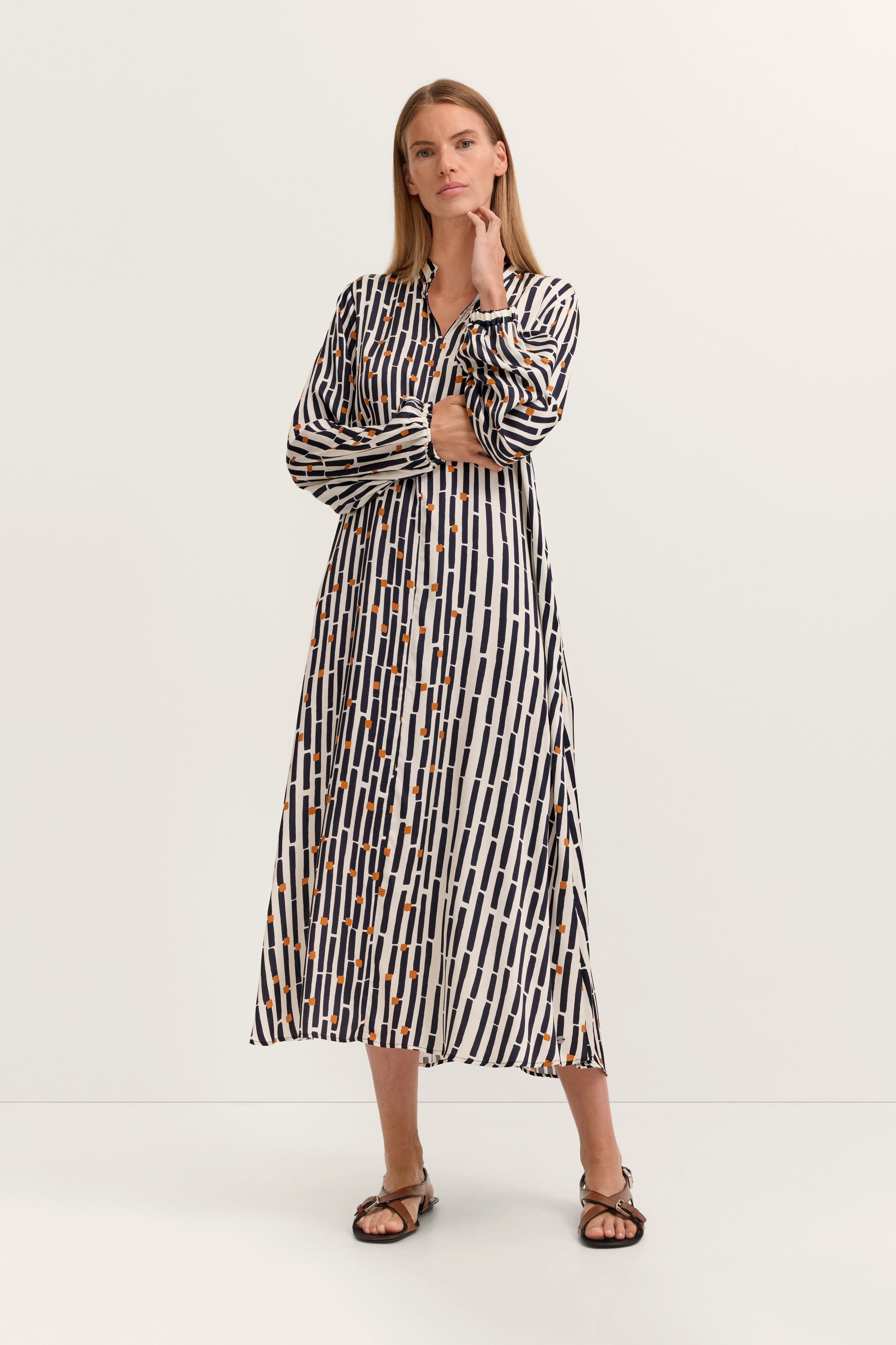 Model in langem Kleid mit grafischem Muster in Blau, Weiß und Orange, Arme verschränkt, entspanntes Posing.