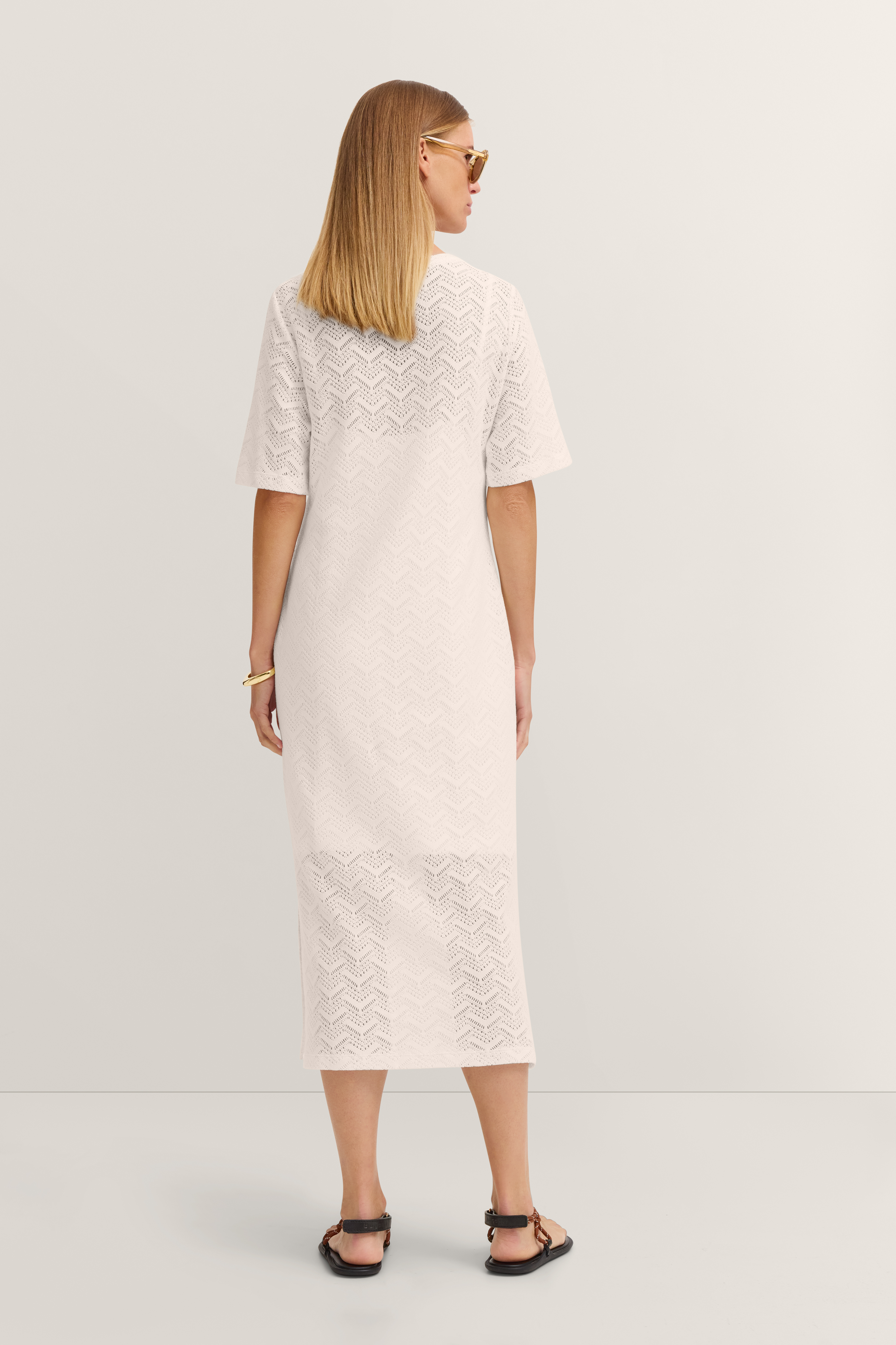 Model in weißem, gehäkeltem bugatti Kleid, entspannt von hinten, mit Sonnenbrille, lässige Pose.