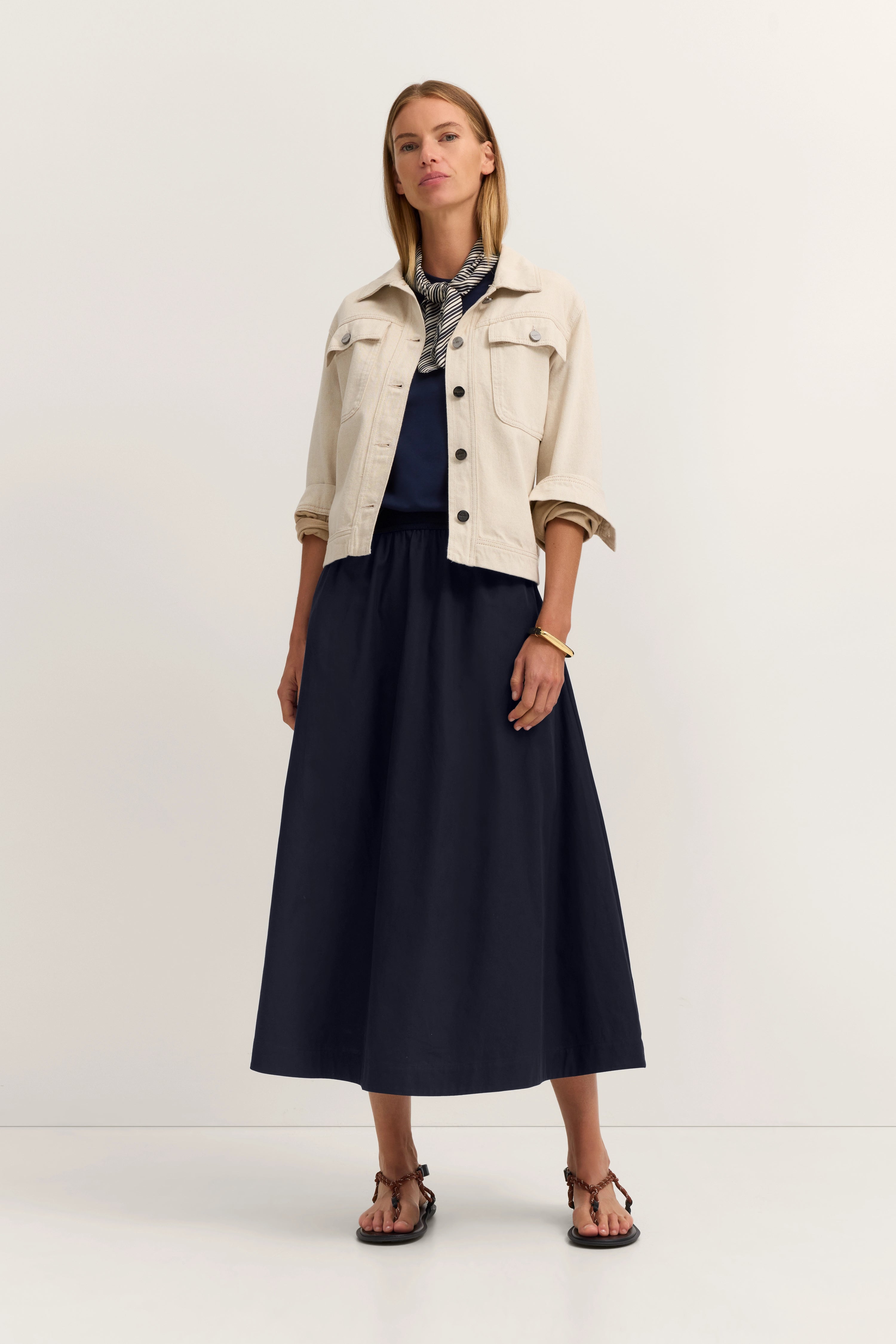 Model in beige Jacke, dunkelblauem Kleid und Sandalen, stehend. Eleganter bugatti-Stil.