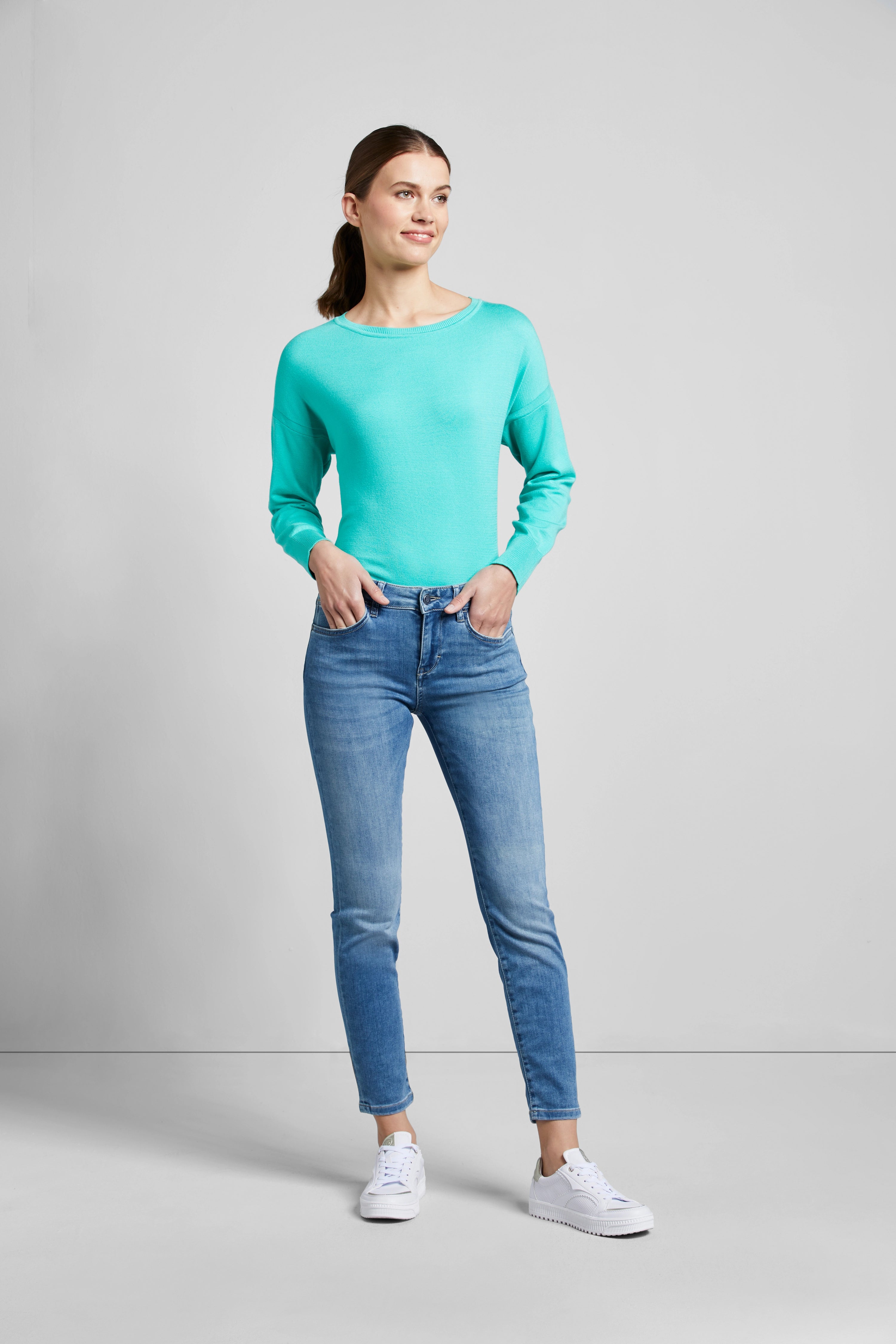 Model in blauwe jeans en groen top, casual pose, witte sneakers.