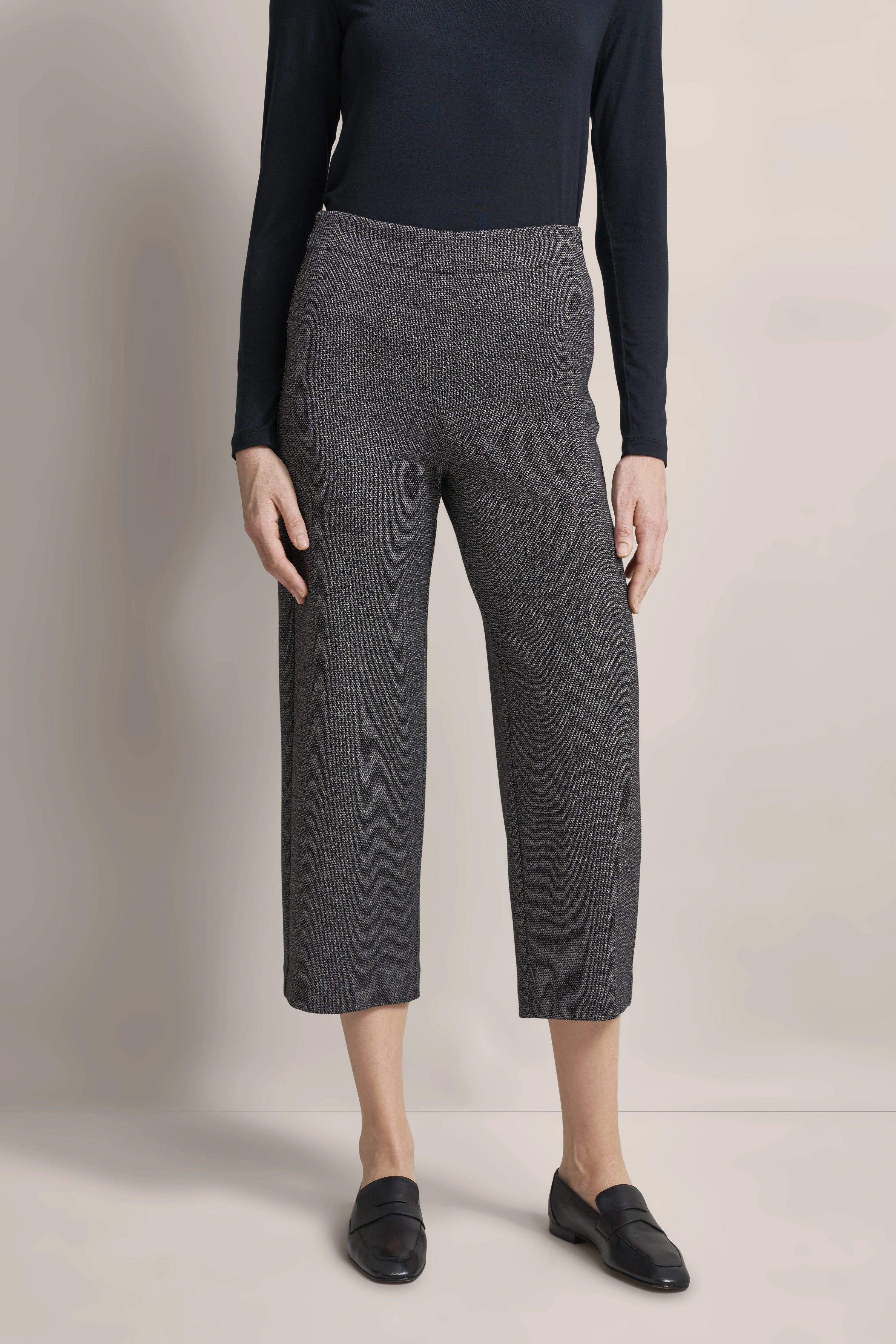 Graue Hose mit Fischgrätmuster, schwarzes Oberteil, Model im legeren Stand, schwarze Loafer.