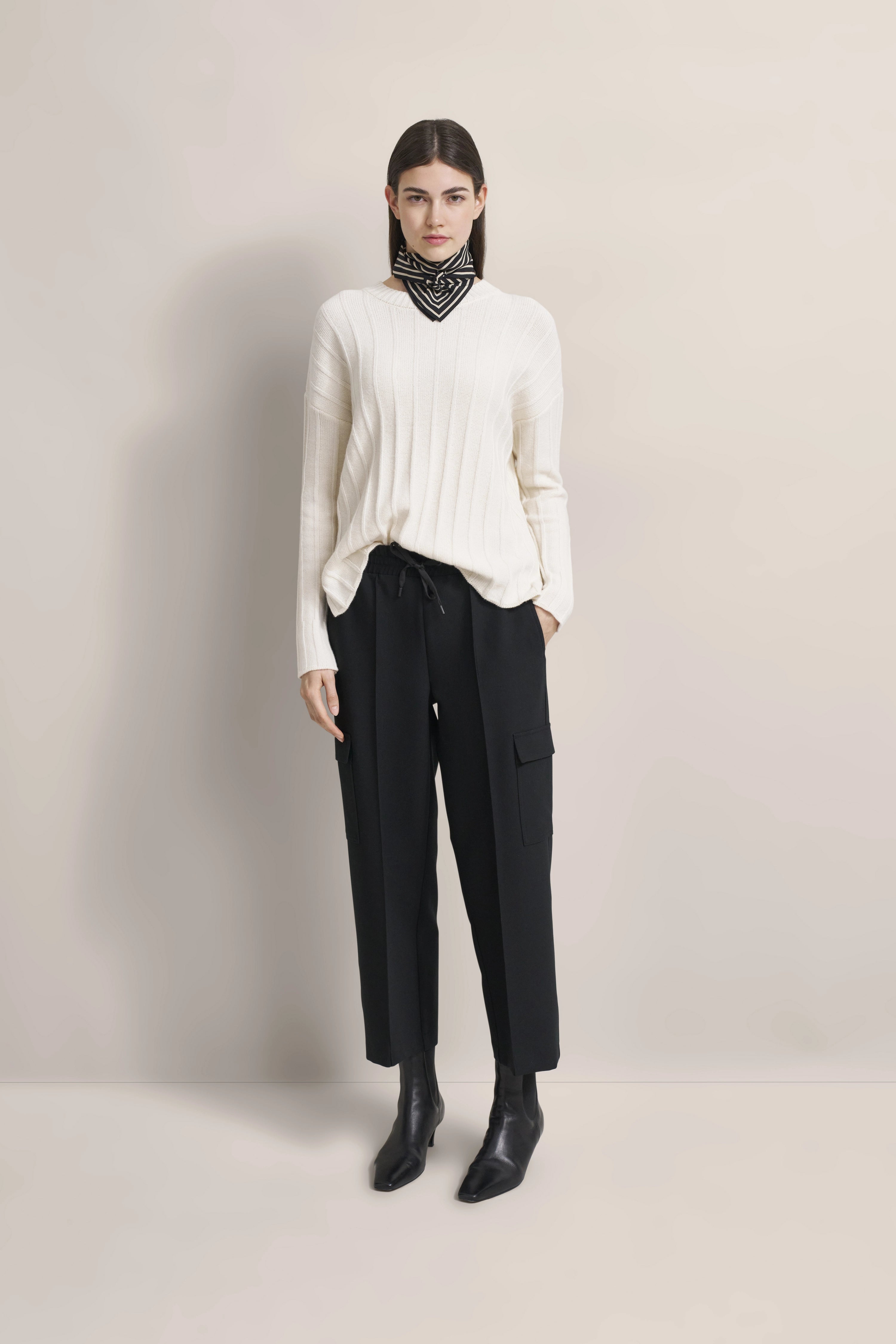 Modelbild: Weißer Pullover, schwarze Hose, schwarze Stiefel. Lockeres Posing, Hände in den Taschen.