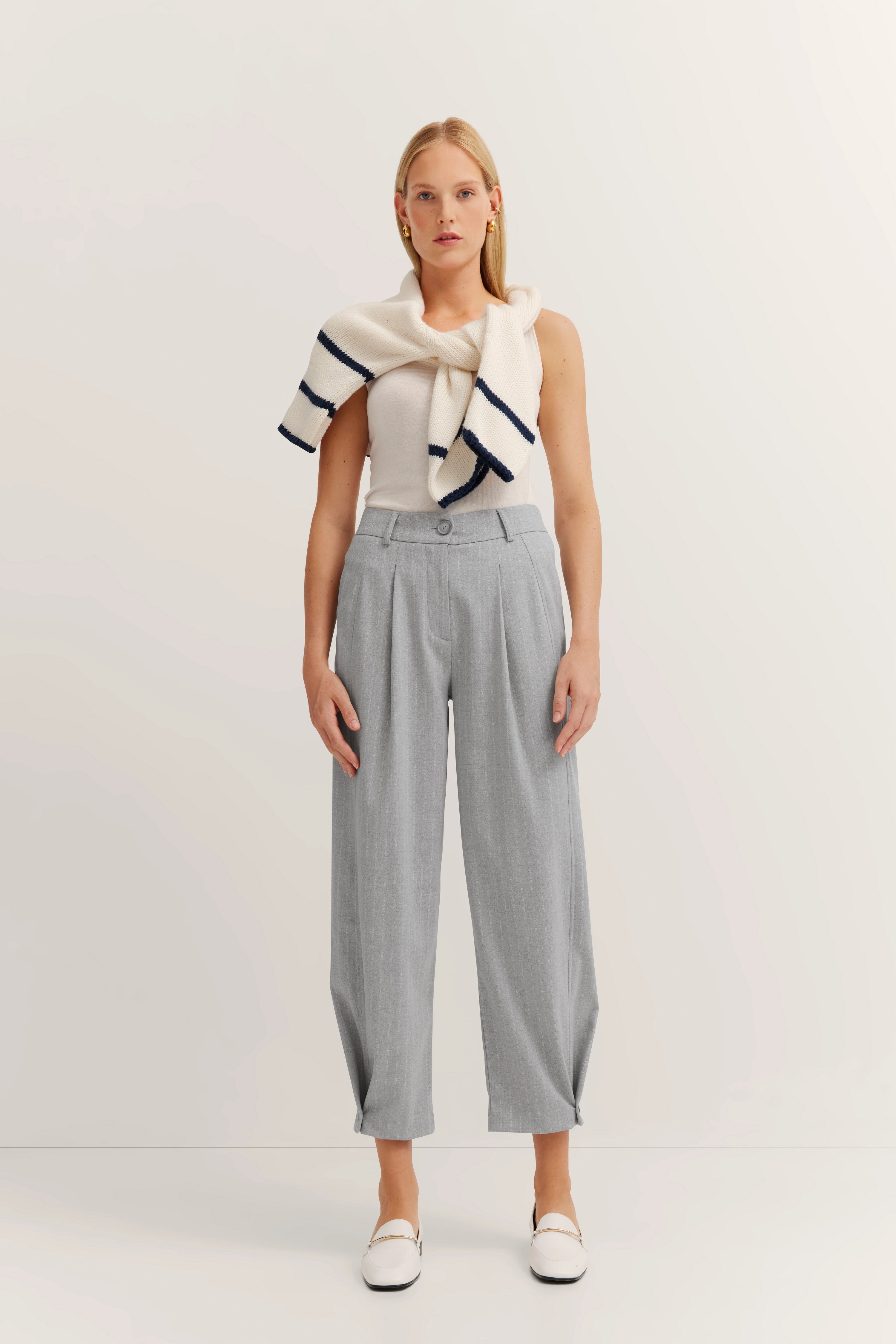 Modelbild: Frau in grauer Hose, weißem Top, Strickpullover über Schulter, elegant und lässig stehend.