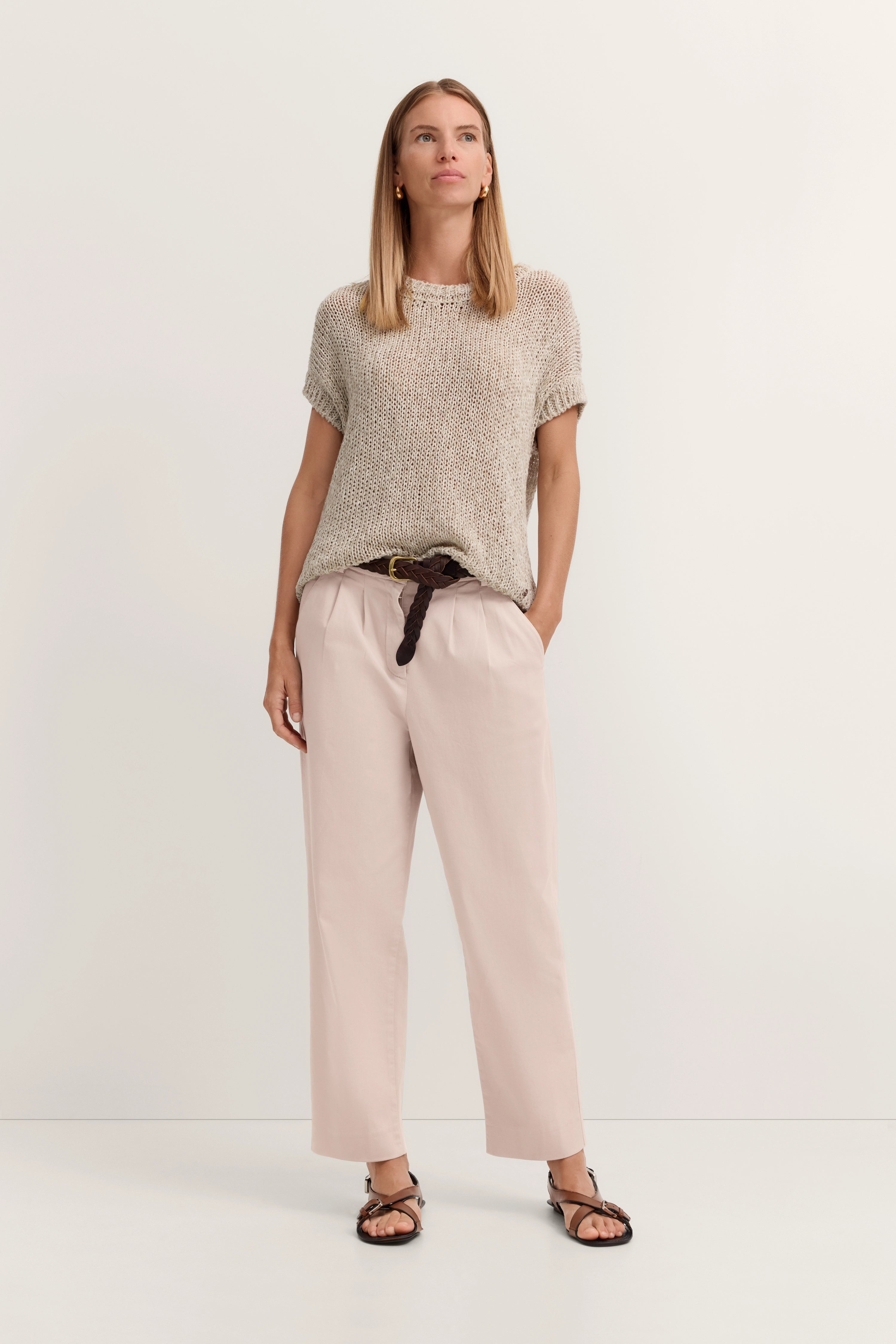 Model in legerem Outfit: beige Stricktop, hellrosa Hose, brauner Flechtgürtel, entspannte Pose.