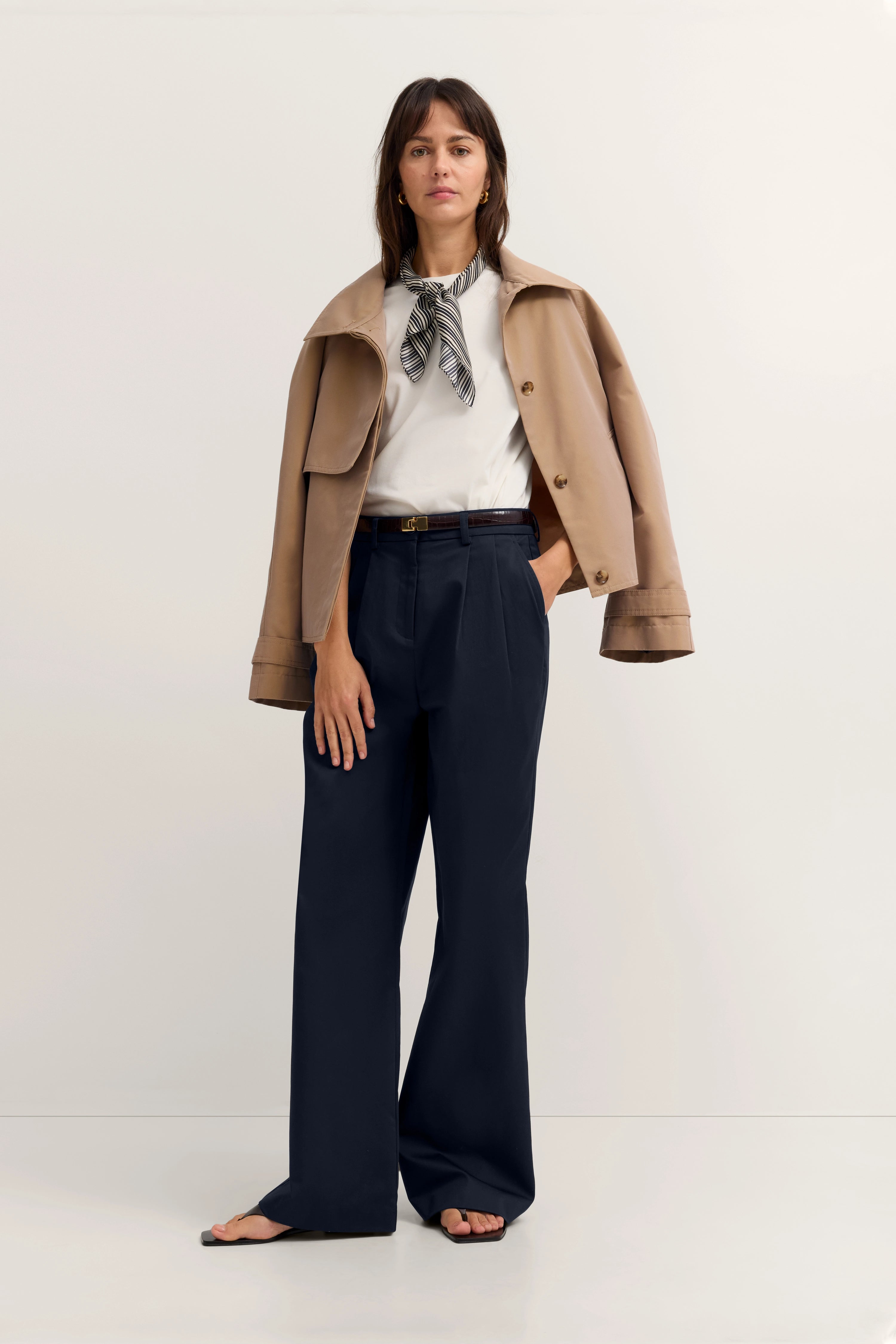 Elegant model in beiger bugatti-Trenchcoat, weißem Top, dunkler Hose und Halstuch, relaxed Pose.