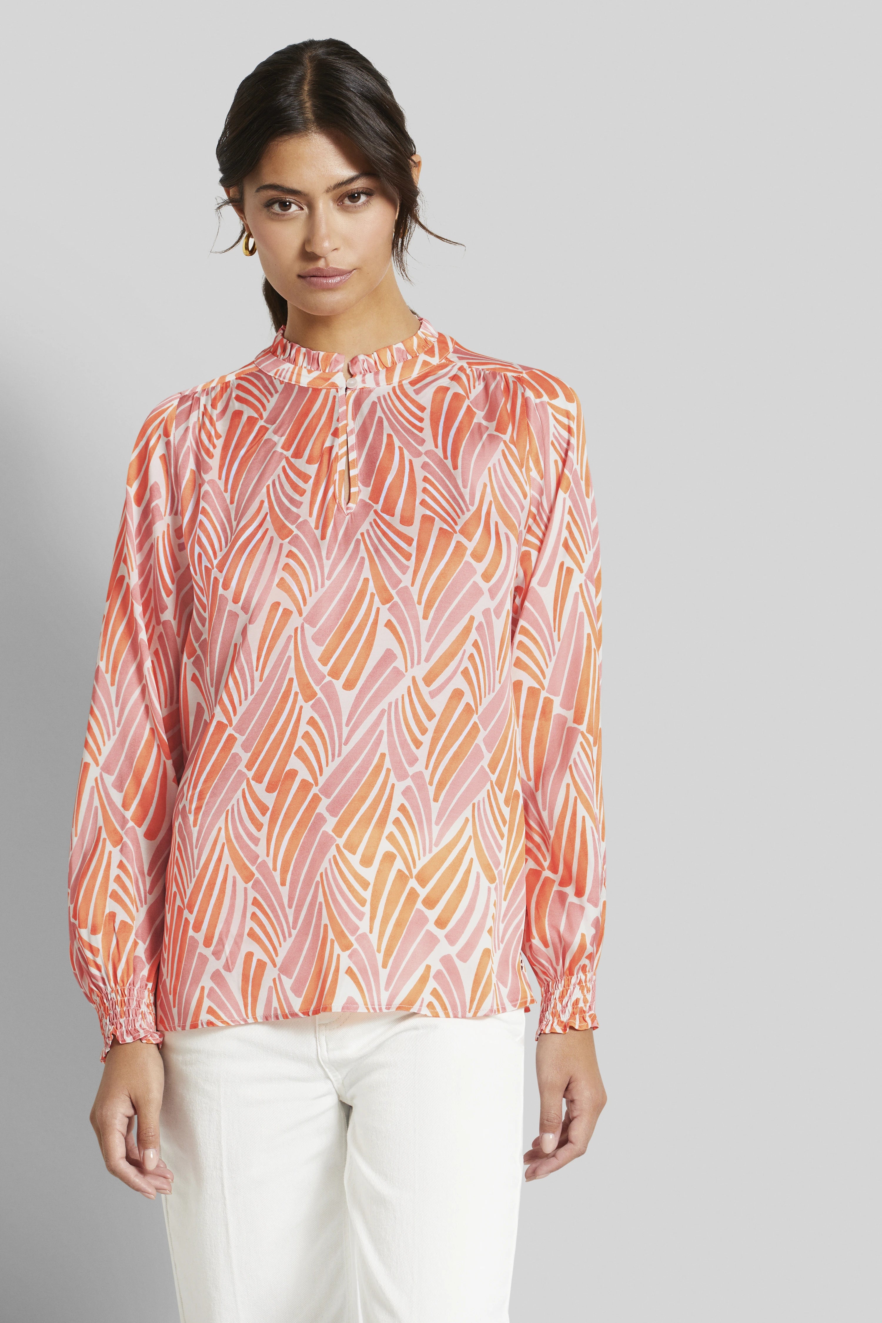 Bluse mit rosa-orange Muster, Modelbild, entspanntes Posing, kombiniert mit weißen Hosen.