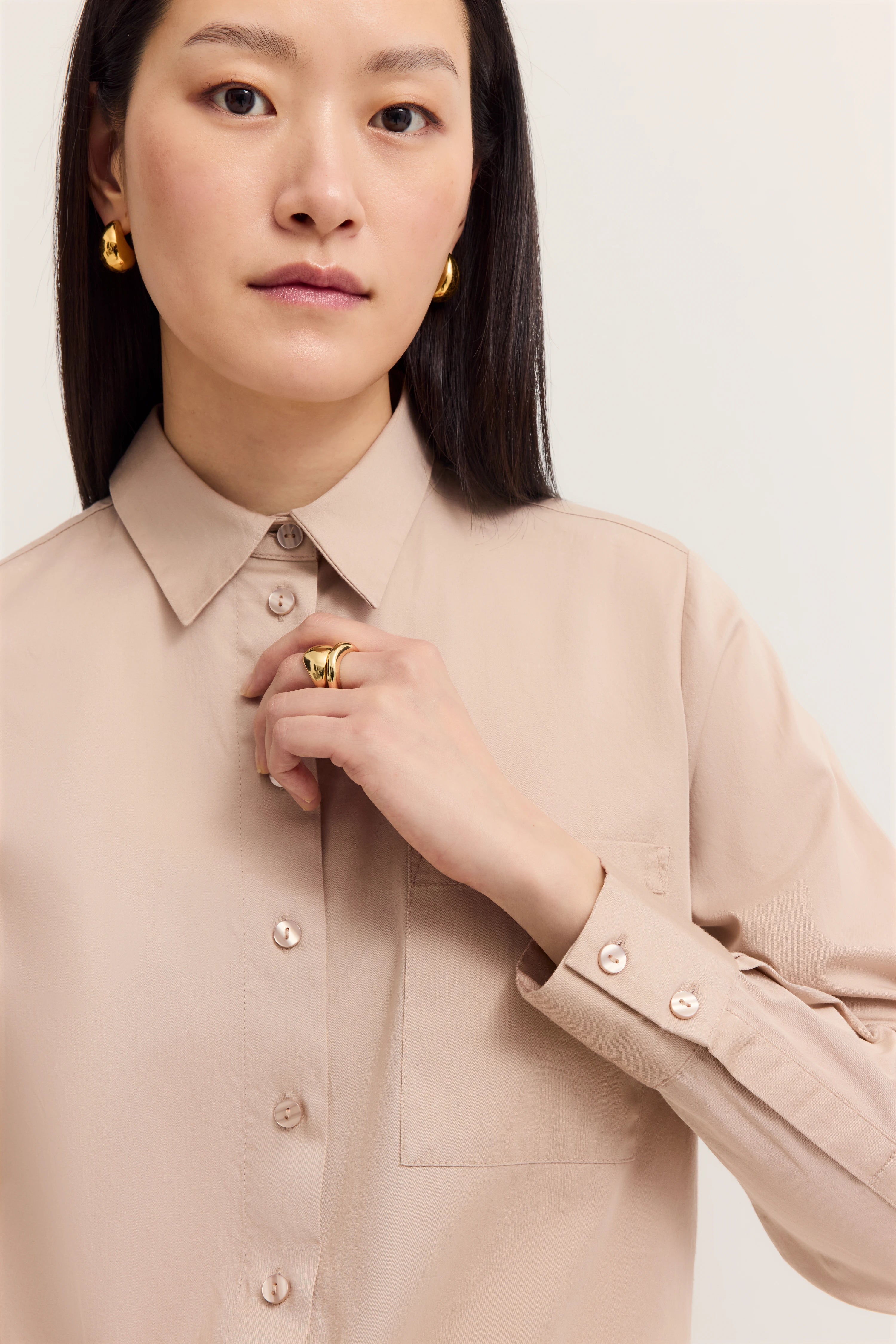 Büstenbild einer Person in beigem Hemd mit goldenen Ohrringen und Ring, elegant und stilvoll.